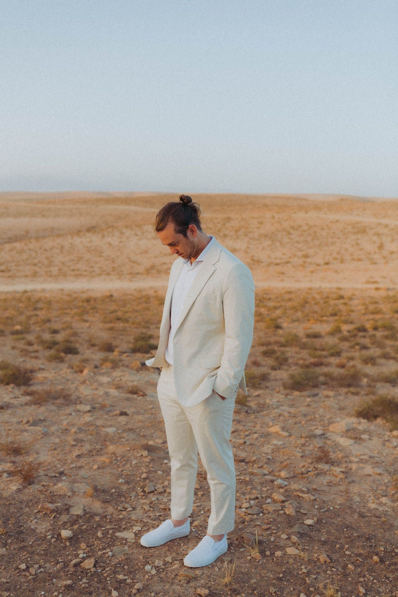 Person wearing an all-white suit and sneakers stands contemplatively in a desert landscape at dusk.