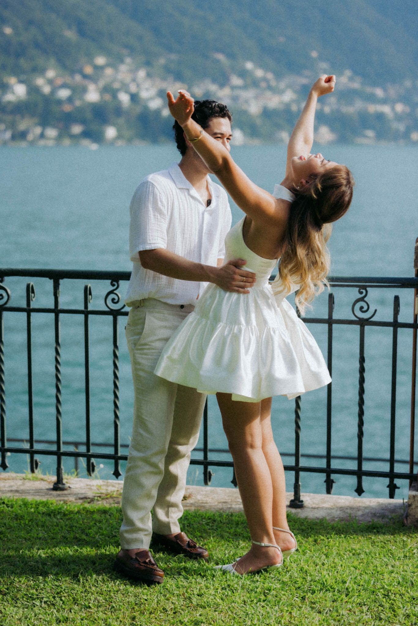 A couple shares a romantic dance moment by a lakeside railing with mountains in the background.
