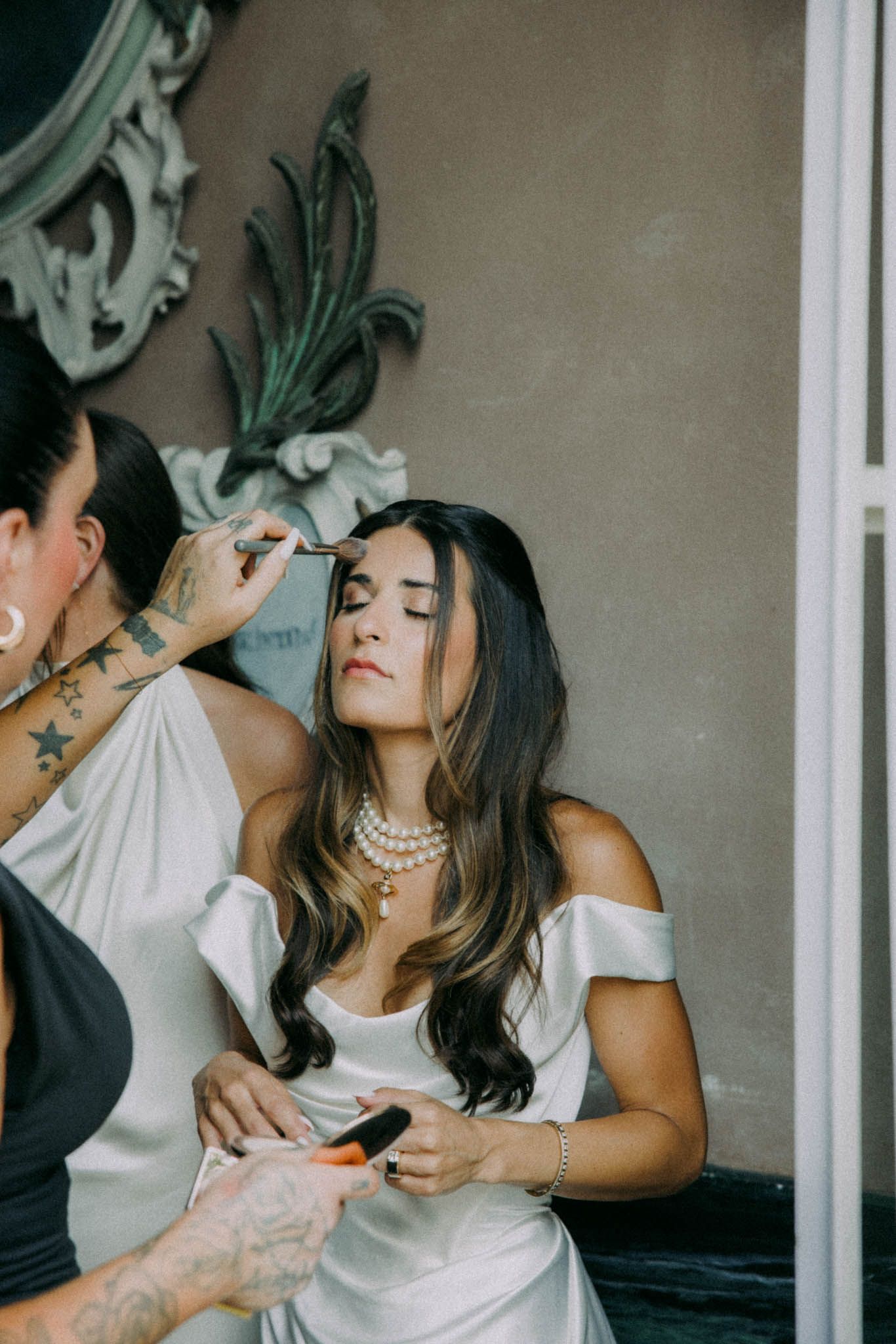 A makeup artist applies cosmetics to a woman in an off-shoulder white dress with pearl necklace during wedding preparations.