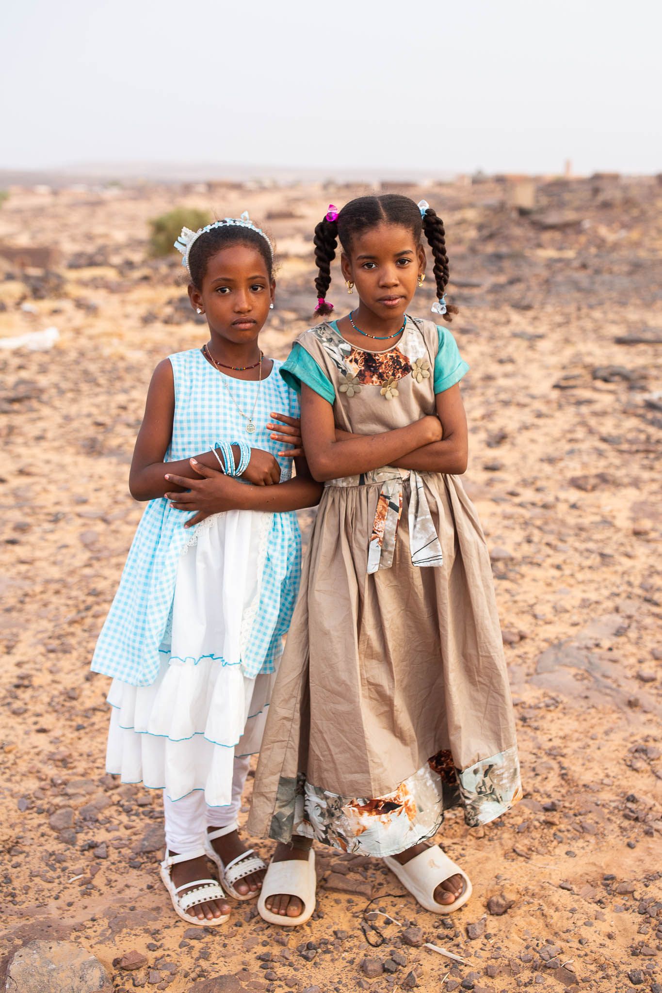 Two young children in dresses stand together on dry desert ground, holding each other in a caring embrace.