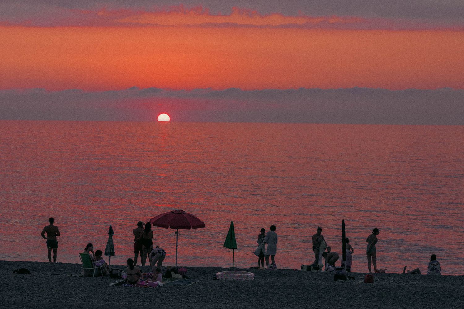 Silhouettes of beachgoers enjoying a vibrant orange and pink sunset over the ocean horizon.