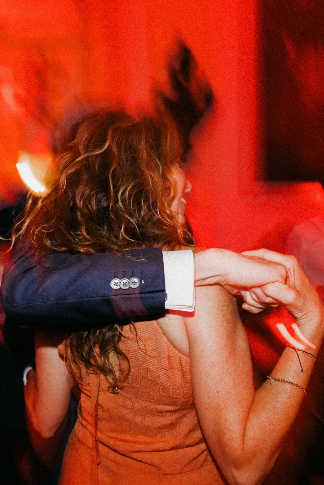 A person in a navy blazer and brown top dances under dramatic red lighting at a party.