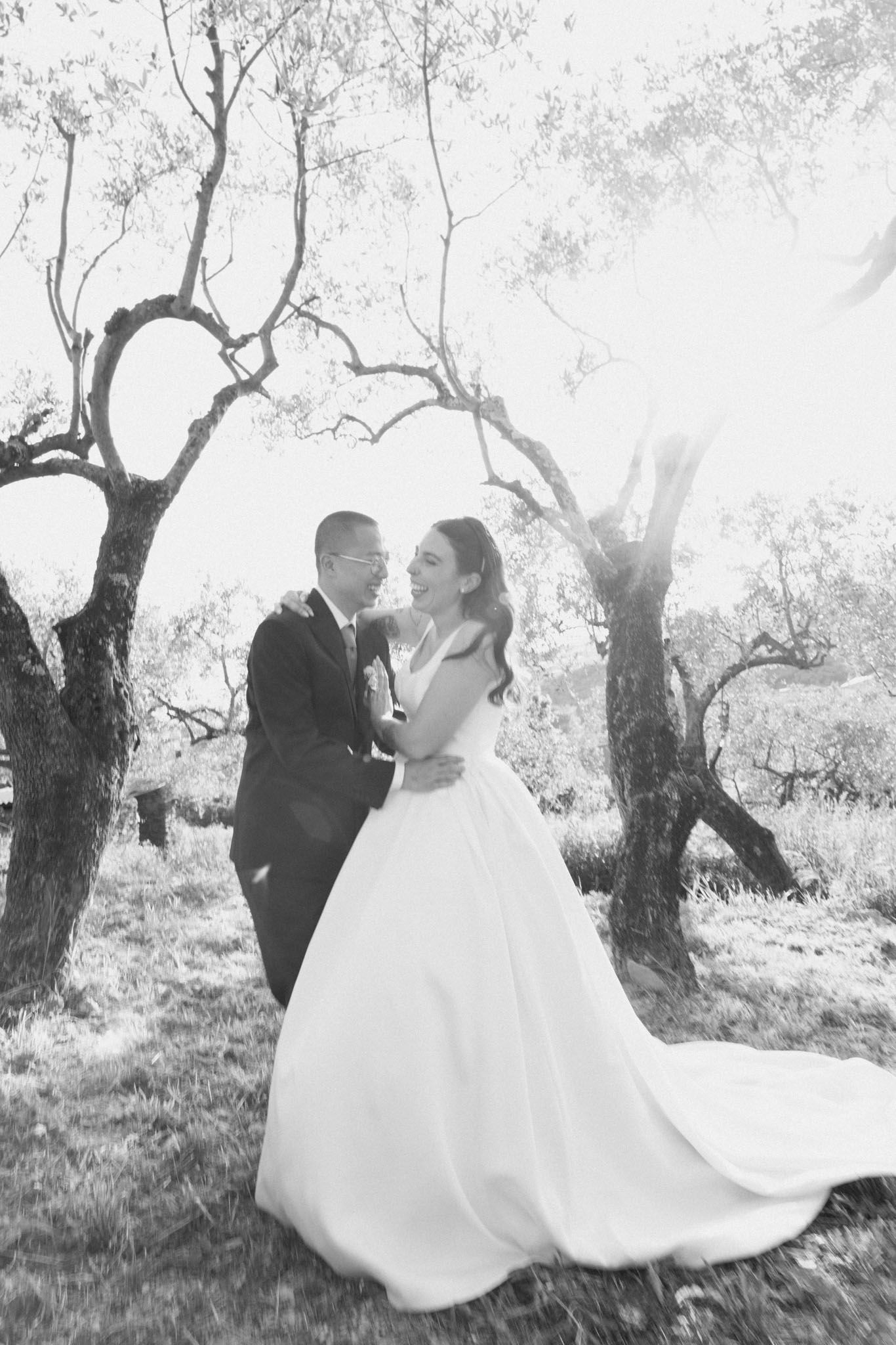 A romantic black and white wedding photo showing a couple embracing under twisted trees in a dreamy outdoor setting.