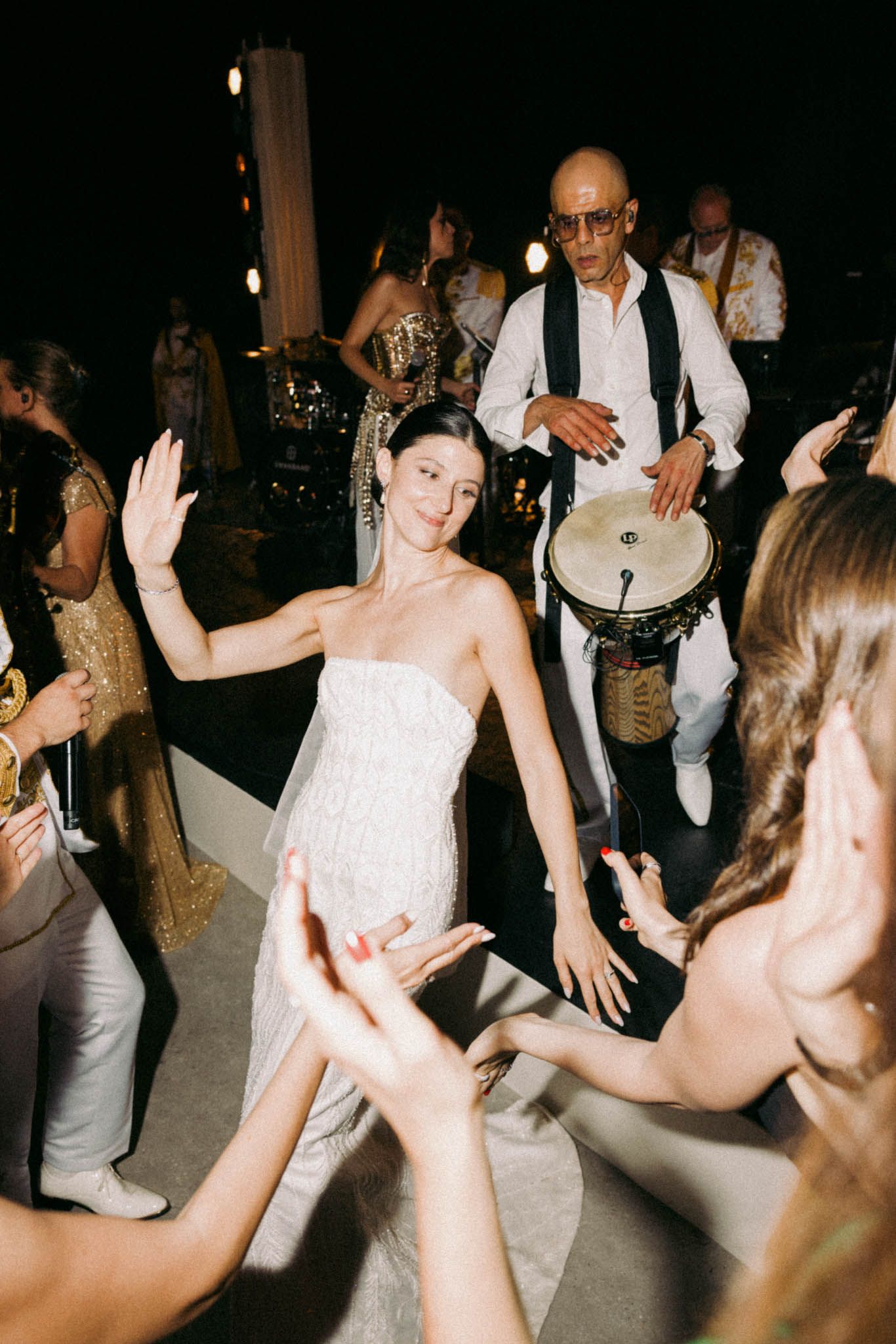 Wedding guests dance energetically on a dark dance floor while musicians perform in the background.