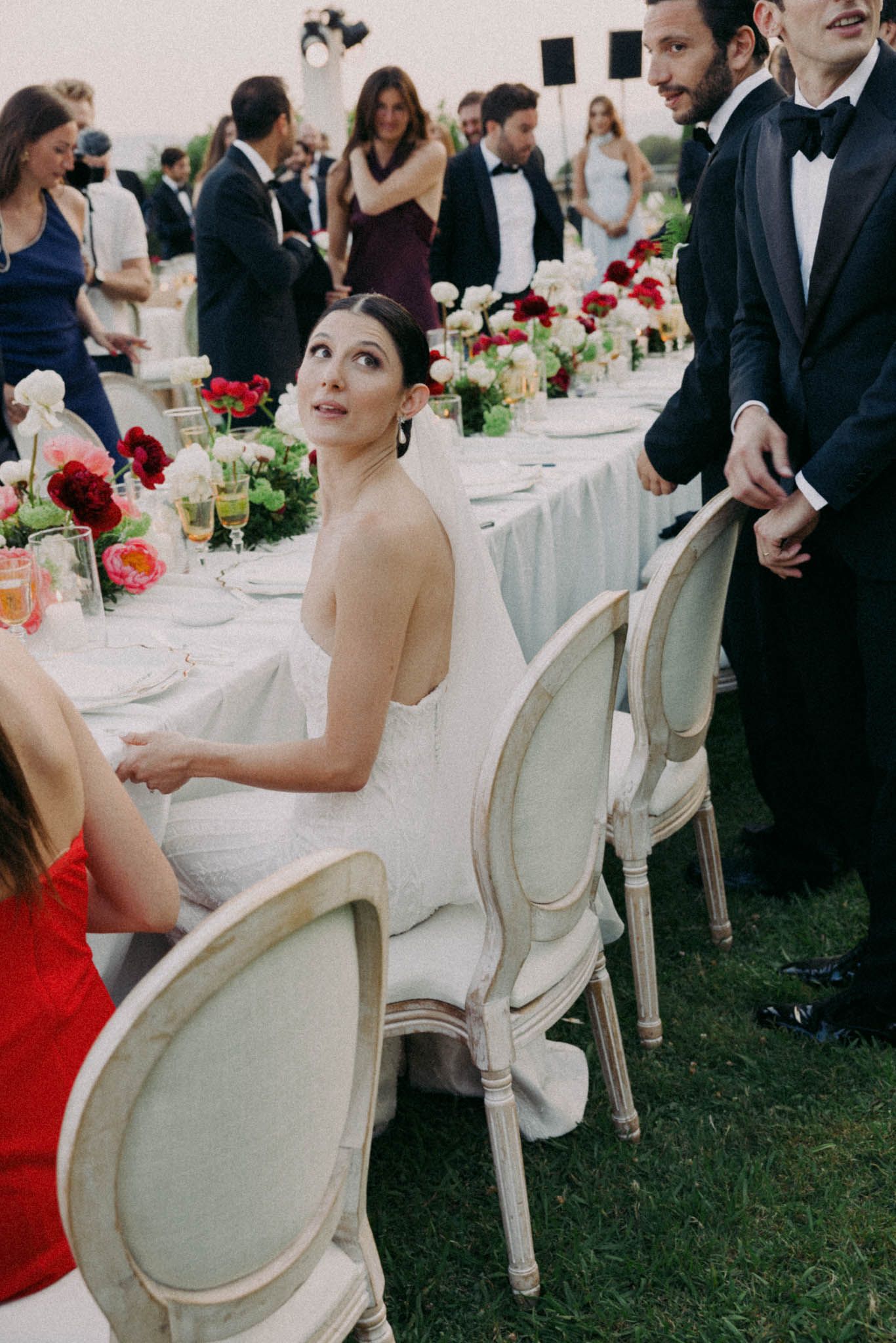 Elegant wedding reception table with red floral centerpieces and guests seated in white French country chairs.