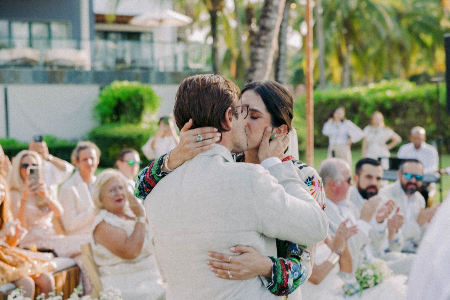 Wedding guests dance and embrace on an outdoor dance floor surrounded by palm trees at a tropical reception venue.