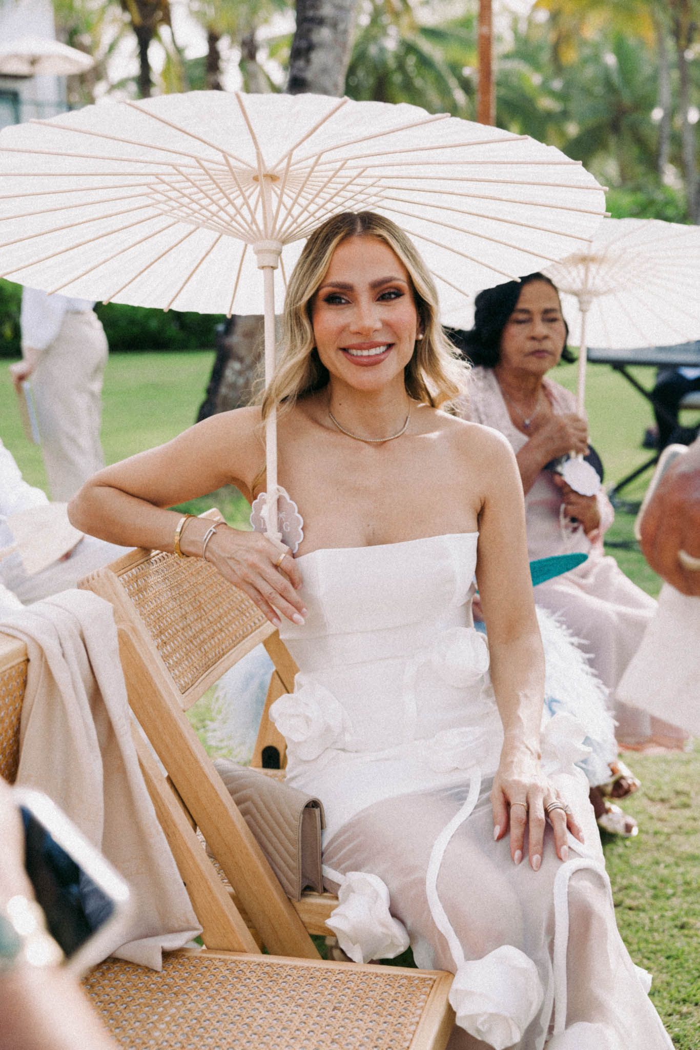 Guests at an outdoor event sit with white parasols in a garden setting wearing elegant white attire.