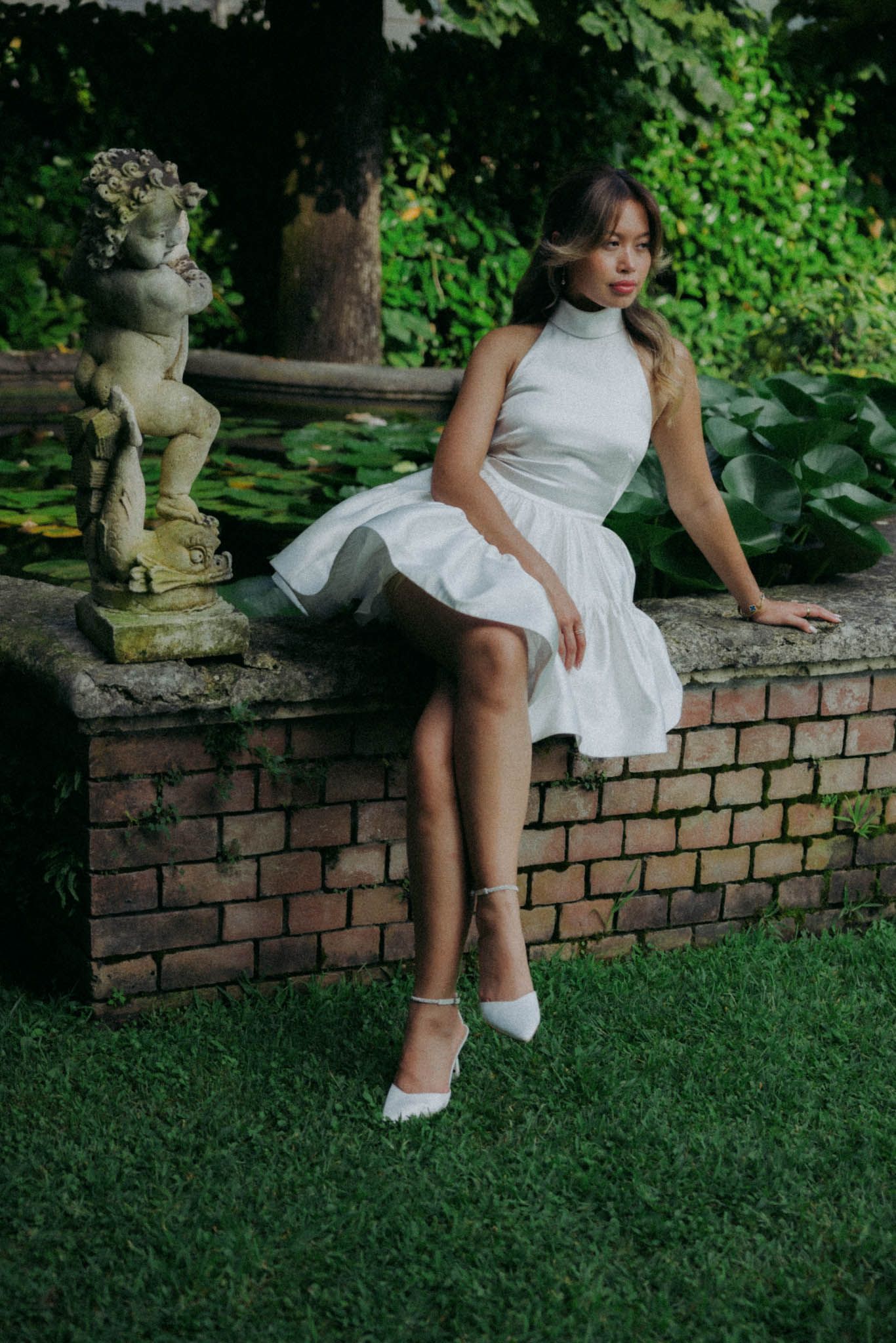 Someone in a white dress sits on a brick wall in a garden setting with lush greenery in the background.