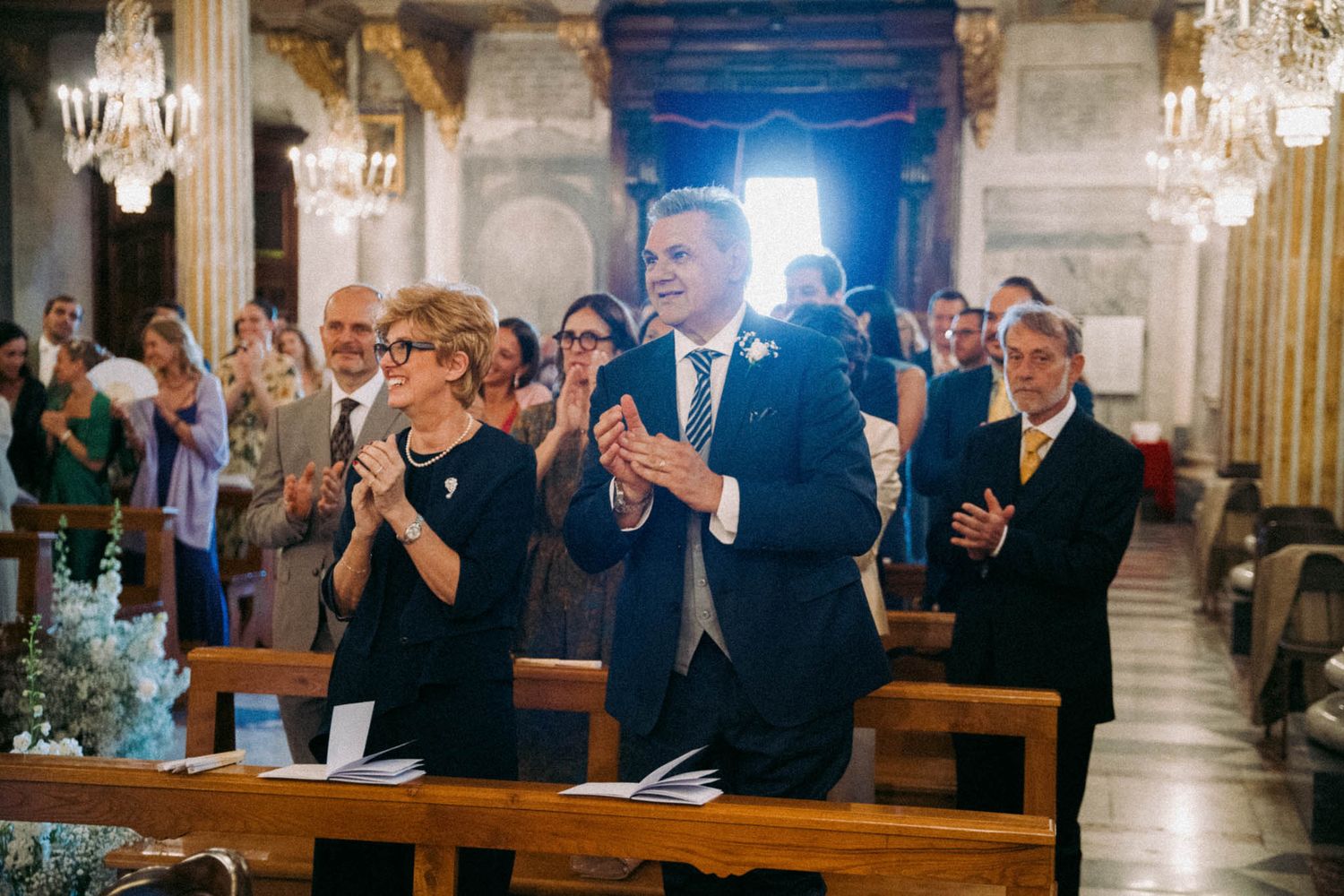 Wedding guests stand and applaud during an elegant church ceremony with crystal chandeliers and ornate architecture.