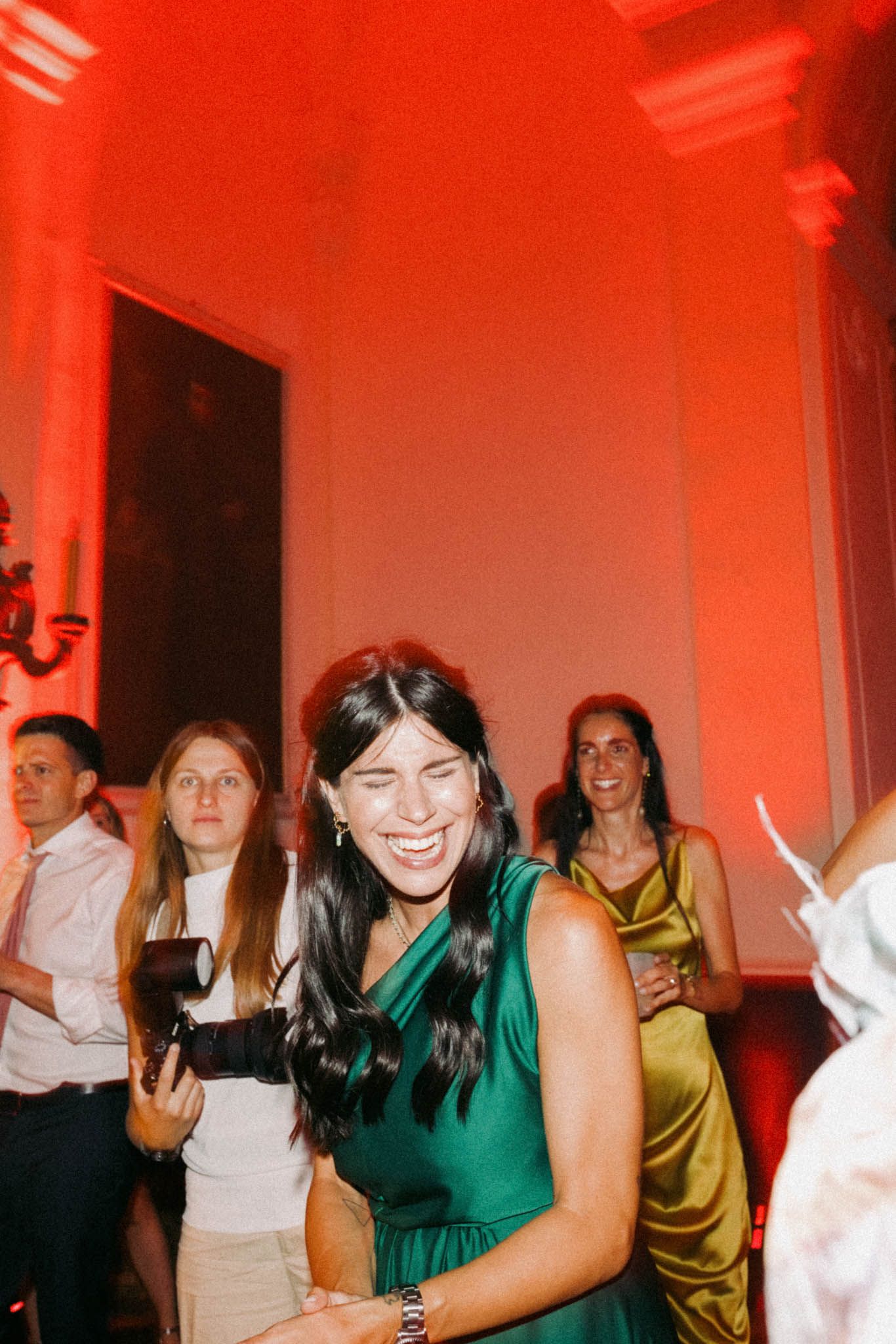 People dancing and socializing at a party with red lighting and white walls.