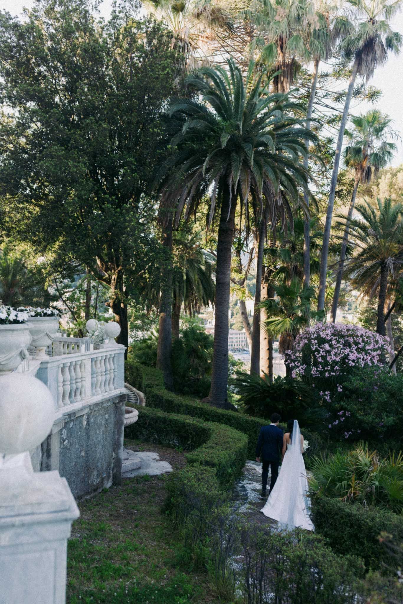 A scenic garden path with white balustrades and palm trees leads down a slope with lush tropical landscaping.