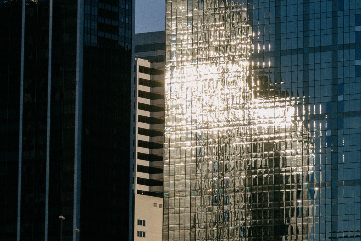 Modern glass office buildings reflect sunlight creating a geometric pattern of light and shadows in an urban setting.