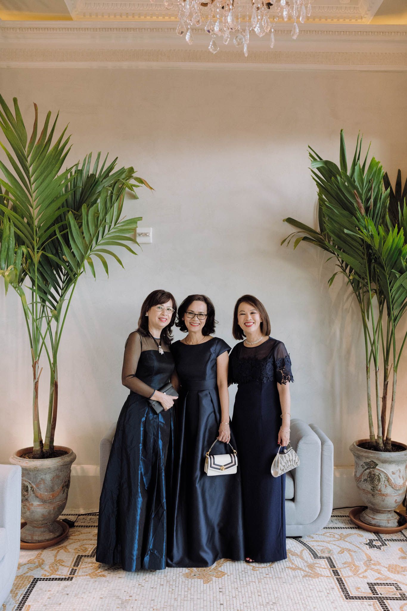 Three women in elegant black dresses stand together in a luxurious room with potted palm plants and ornate decor.