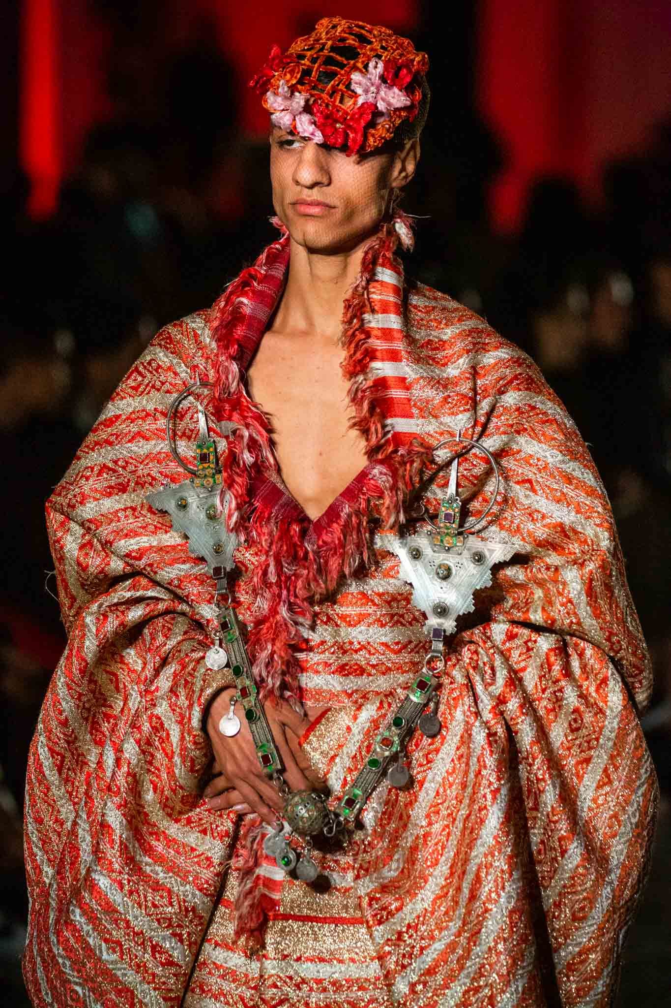 Fashion model wearing red and white striped draped fabric with matching headpiece on runway during fashion show.