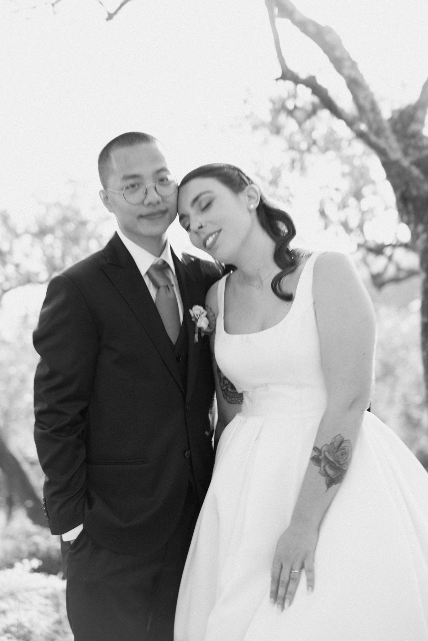 A romantic black and white wedding photo of a couple embracing outdoors under a tree.