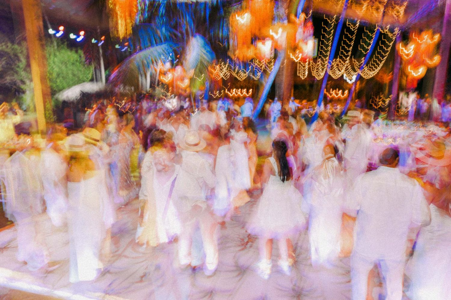Motion blurred crowd of people in white attire dancing under colorful lights and decorations at a nighttime celebration.
