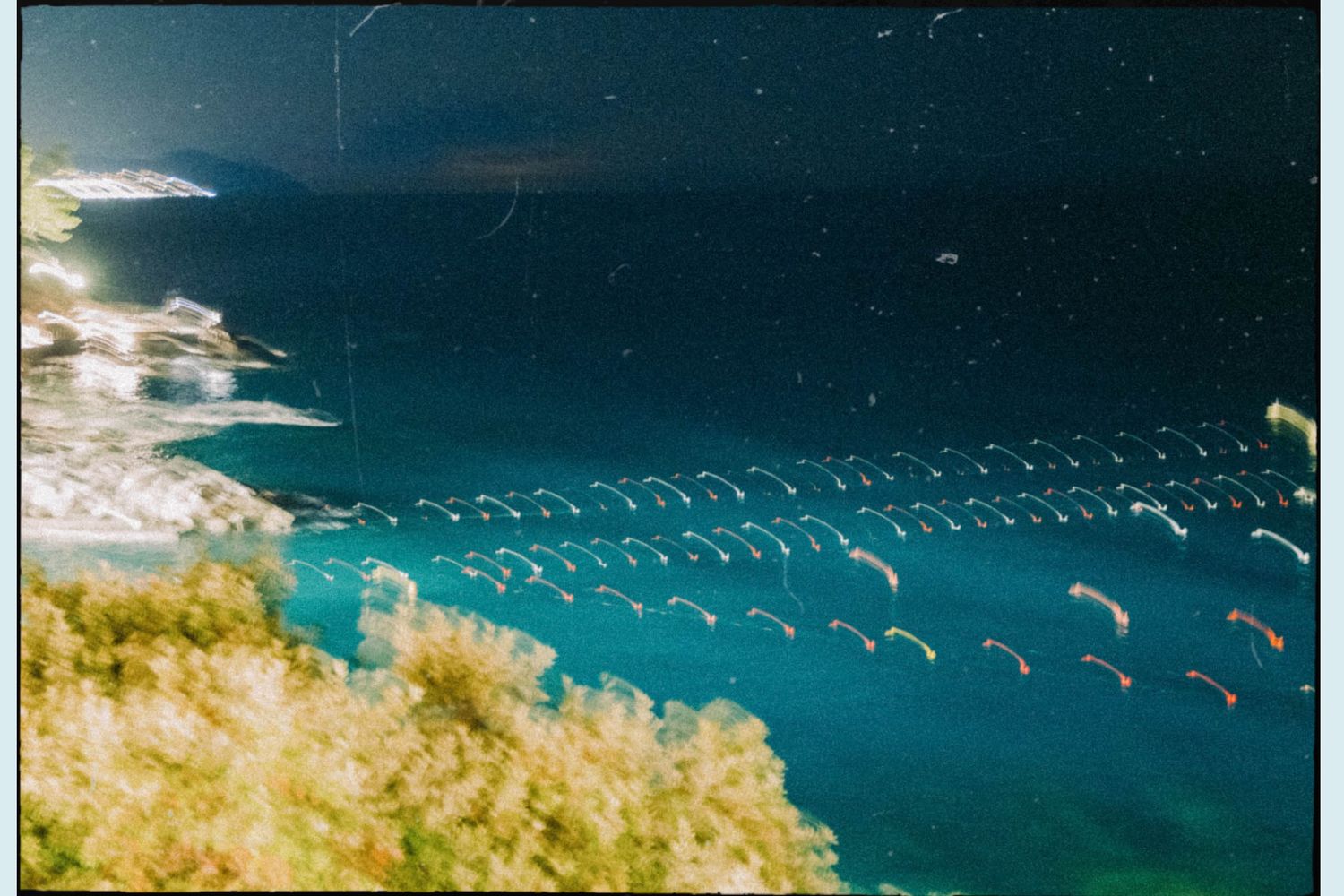 Aerial view of turquoise ocean waters with floating buoy lines and coastal vegetation visible in bright sunlight.