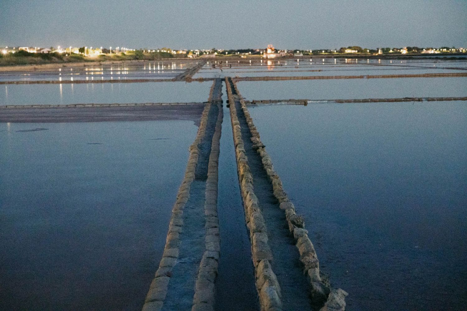Two parallel pipelines extend across calm blue water at sunset with coastal buildings visible in the distance.