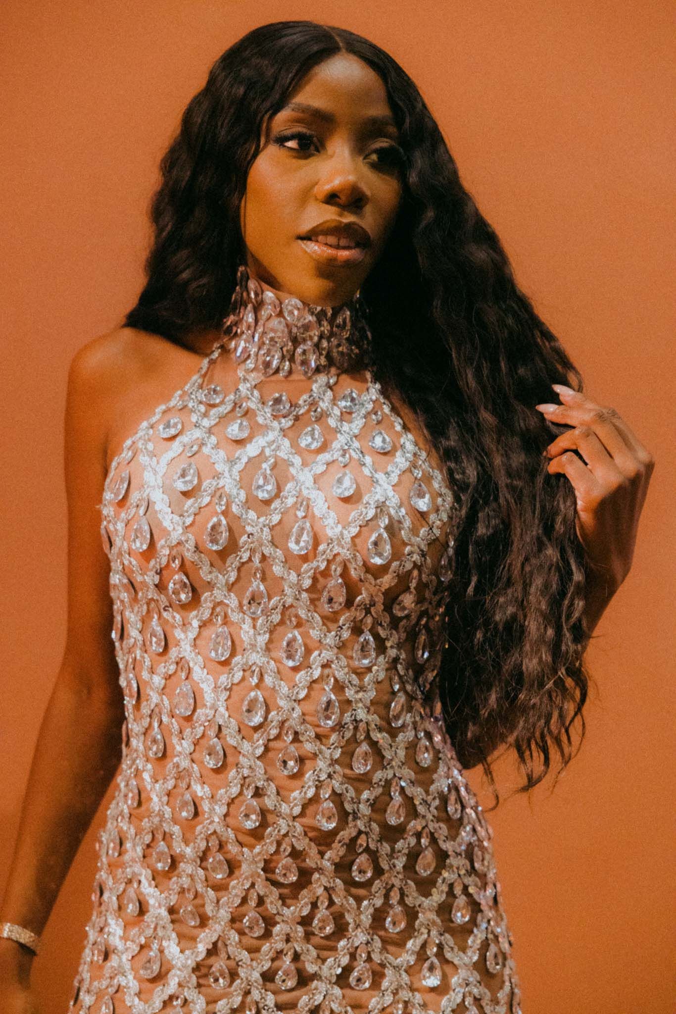 Portrait against orange background wearing a high-neck beaded gown with intricate lace pattern and flowing long dark hair.