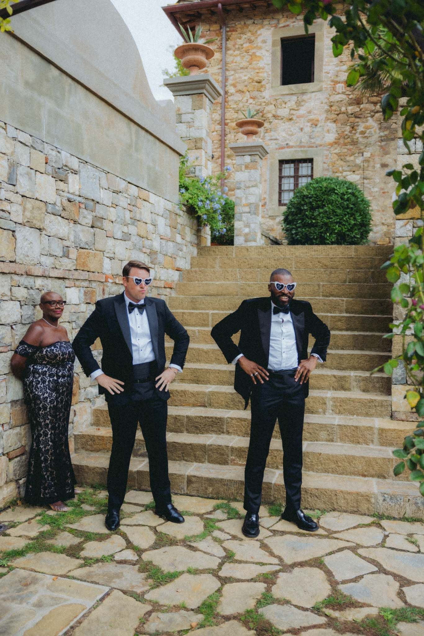 Three people in formal black tie attire pose on stone steps outside a rustic Mediterranean villa.