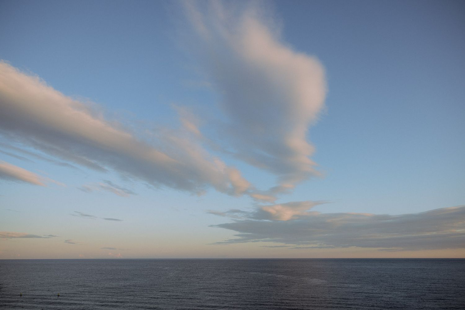 Wispy pink clouds drift across a blue sky over a calm ocean horizon at dusk.