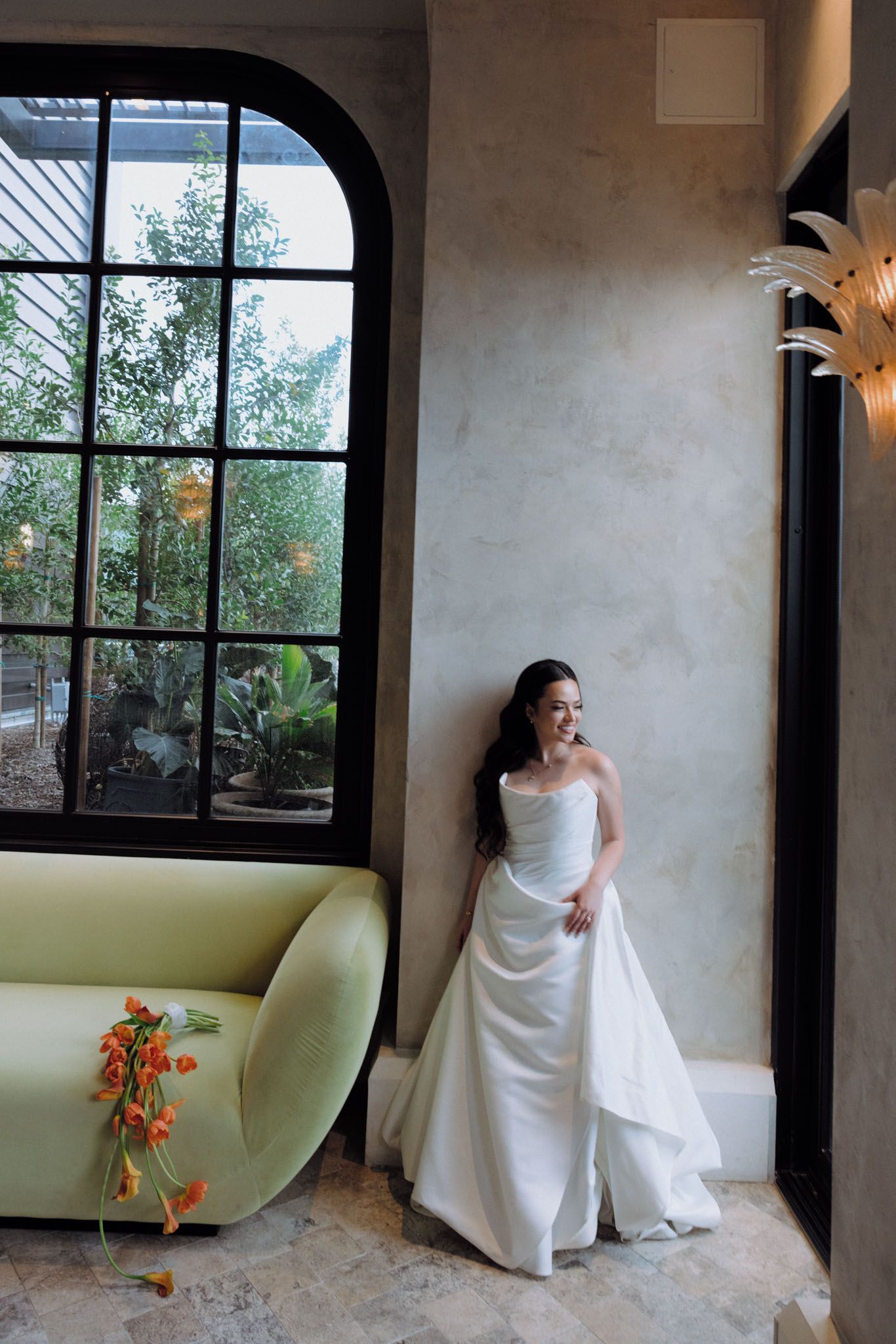 A bride in a white wedding dress poses against textured walls and black-framed windows in a rustic indoor setting.