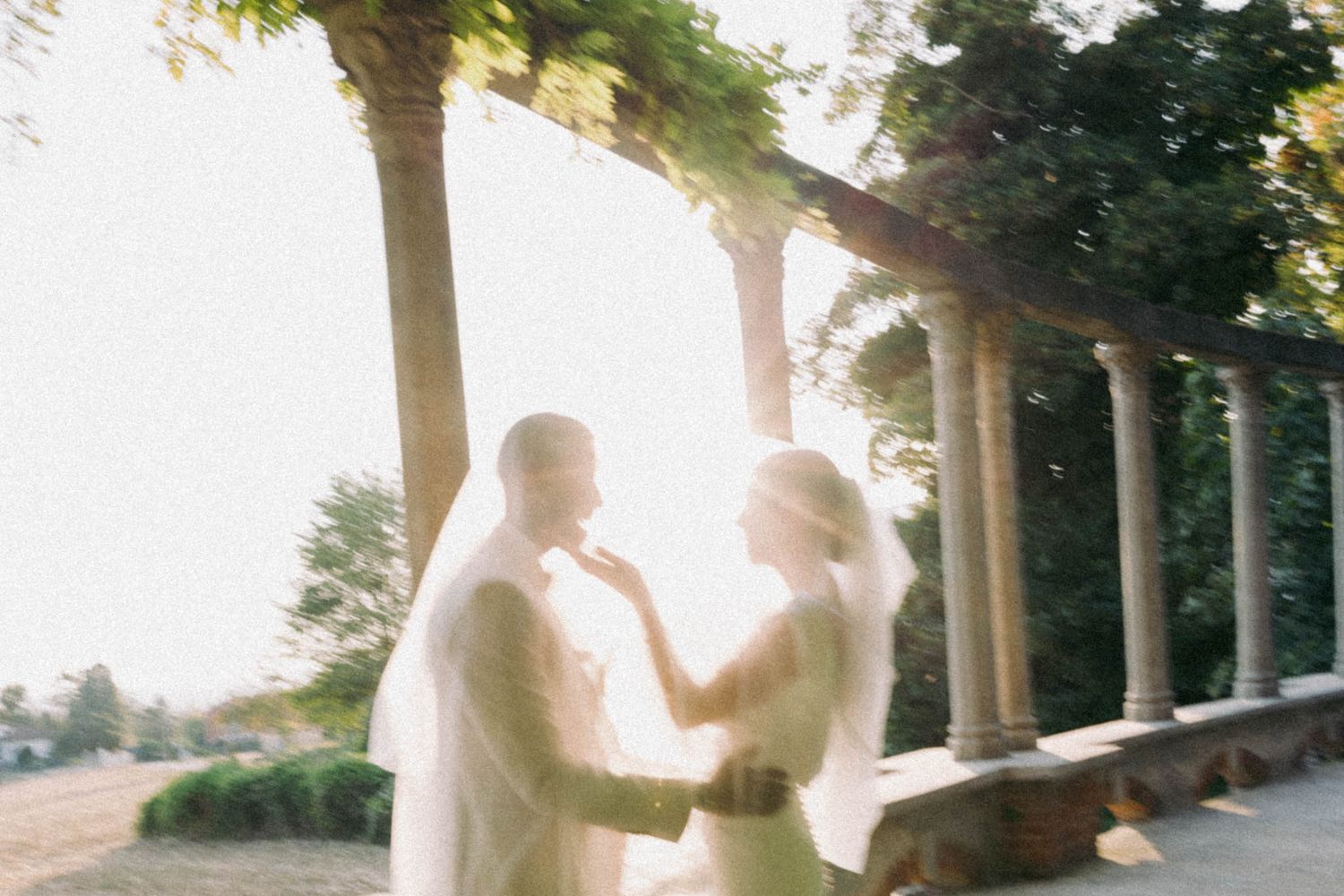 A couple embraces in a romantic outdoor setting with sunlight filtering through trees along a columned walkway.