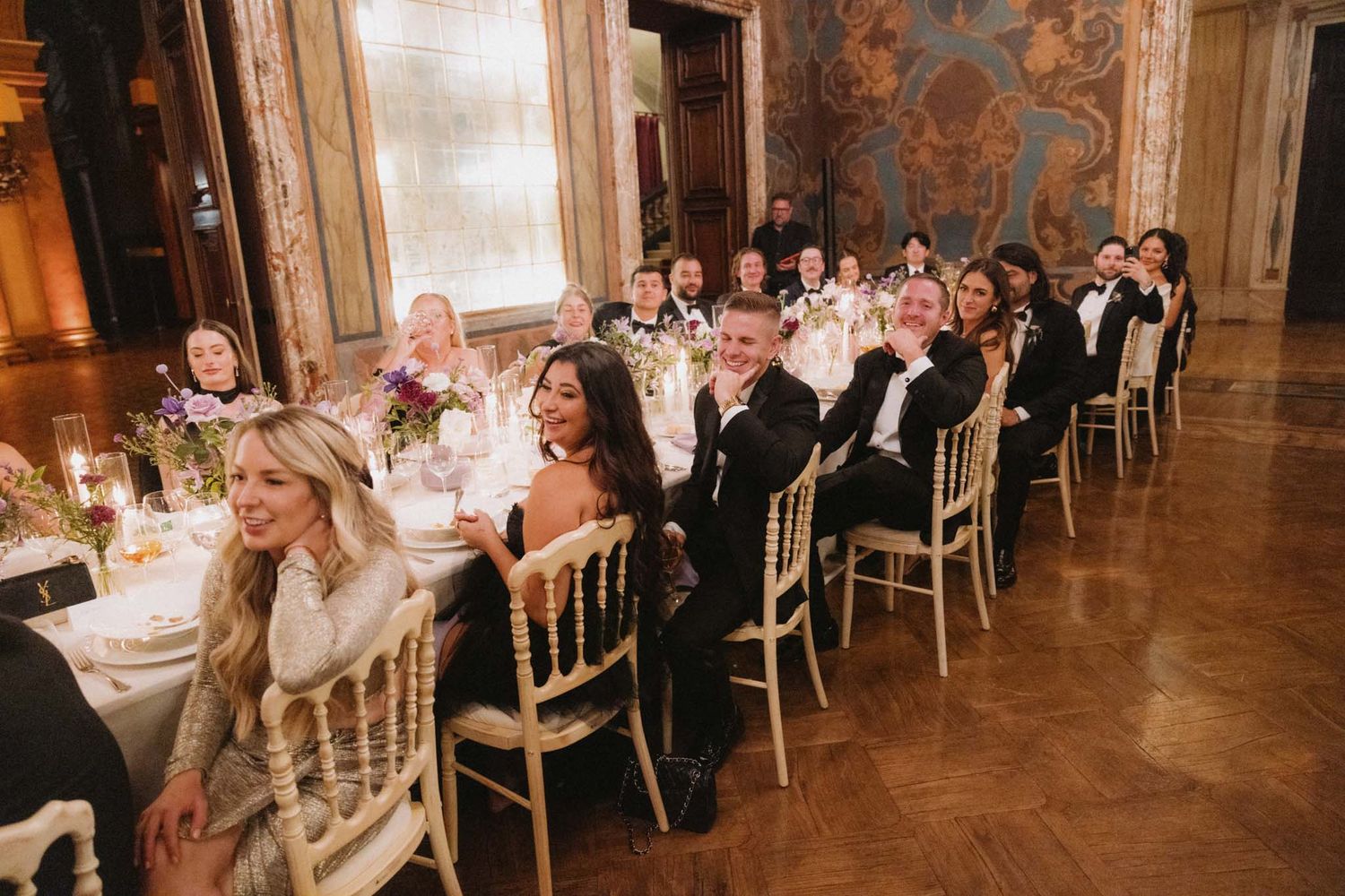 Wedding reception guests seated at elegant round tables in a grand ballroom with ornate decor and warm lighting.