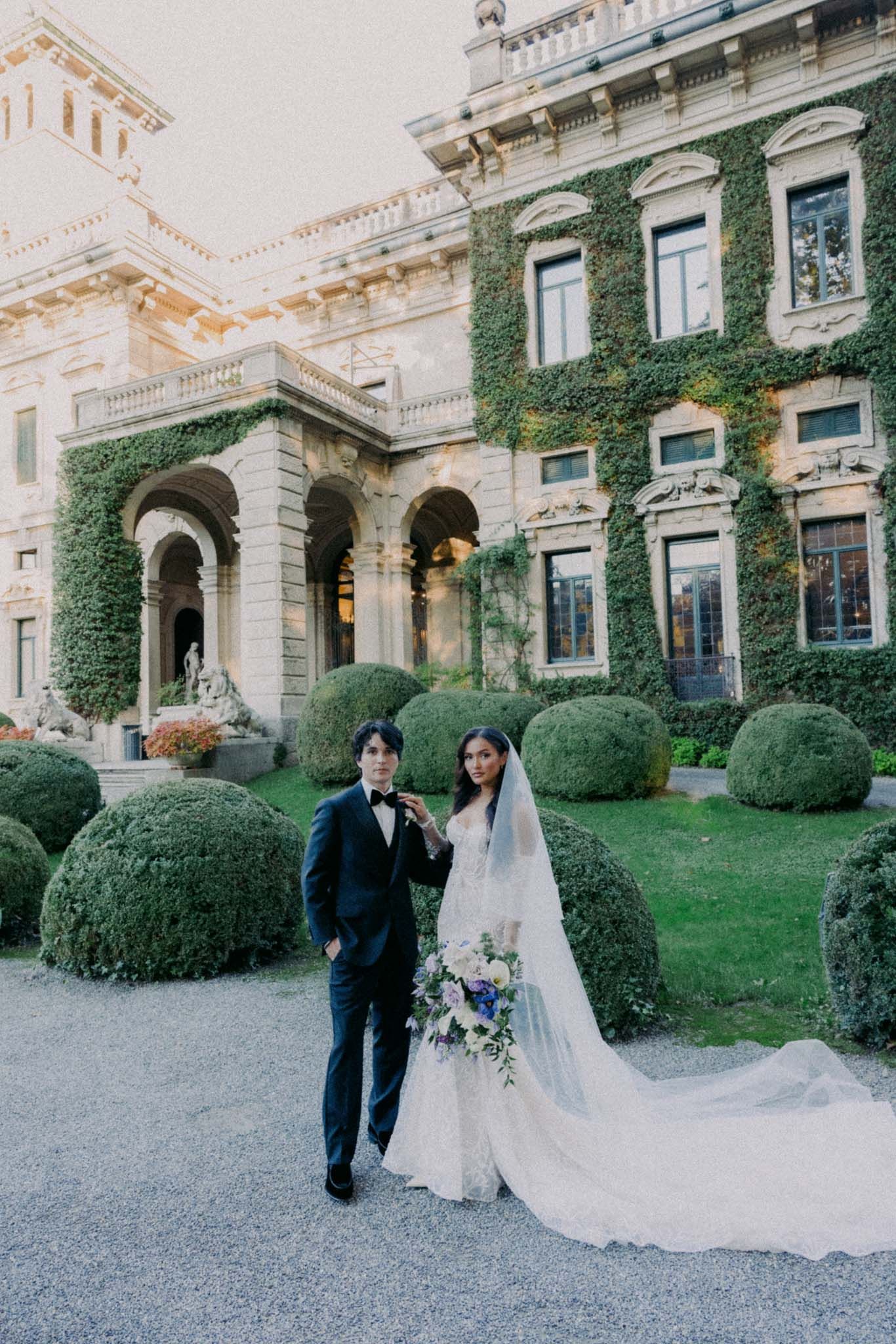 Newlyweds walk together on gravel path beside historic stone building with climbing vines.