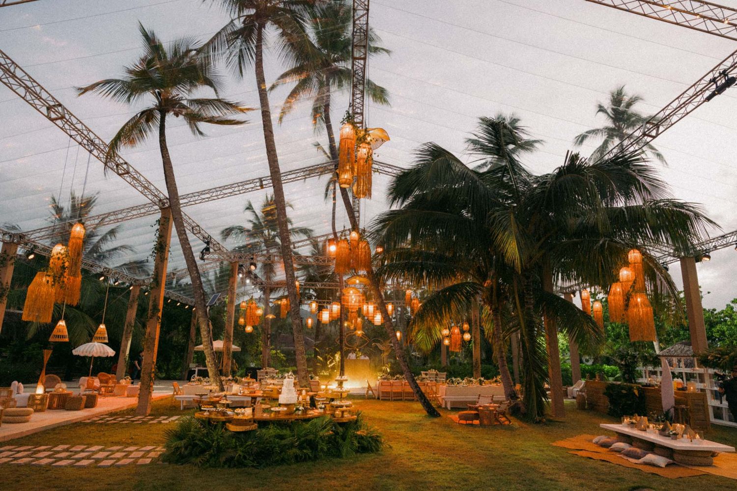 Panoramic view of palm trees and glowing lanterns along a tropical beach at sunset.
