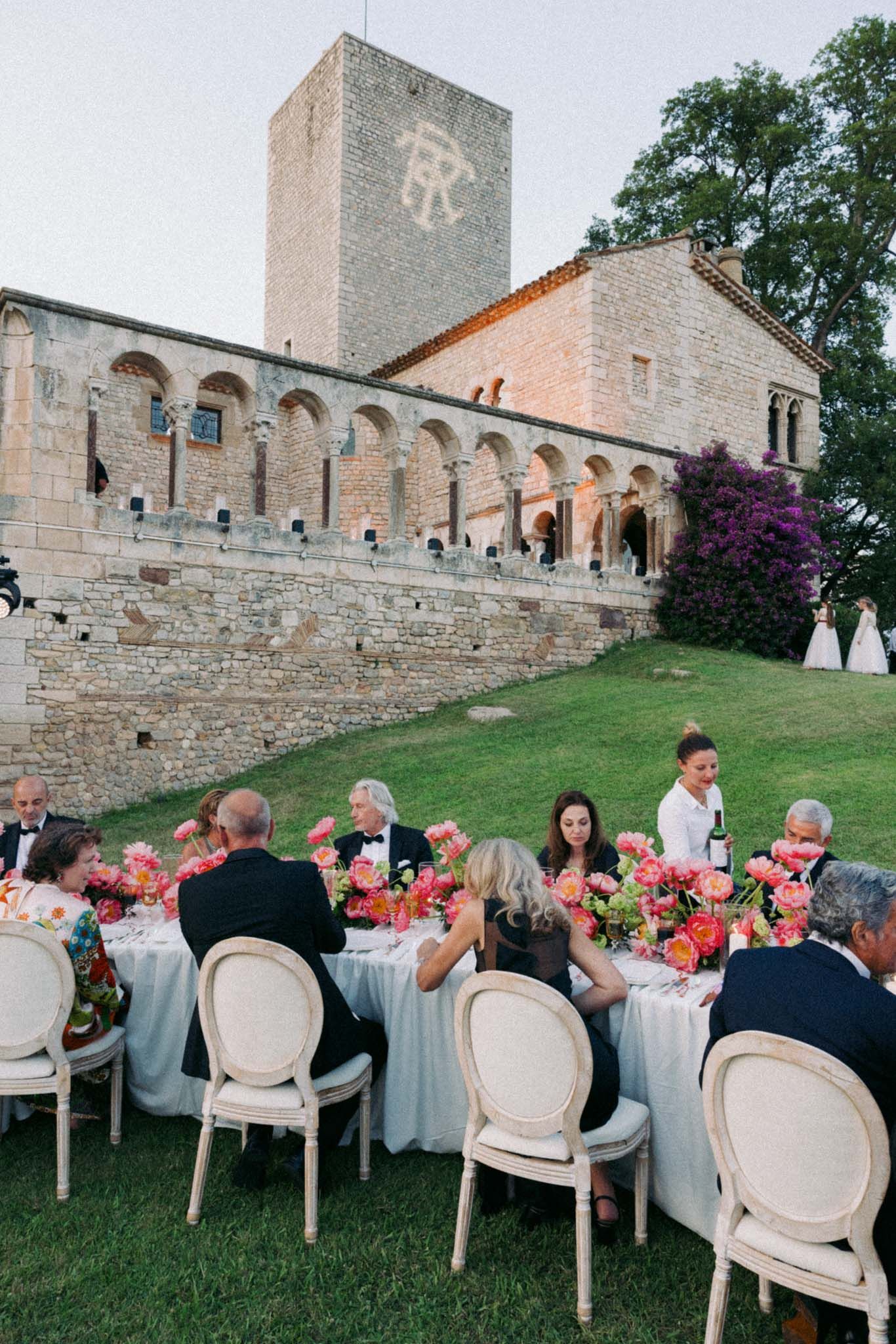 Outdoor wedding reception setup with elegant white chairs and floral centerpieces along stone fortress walls at sunset.