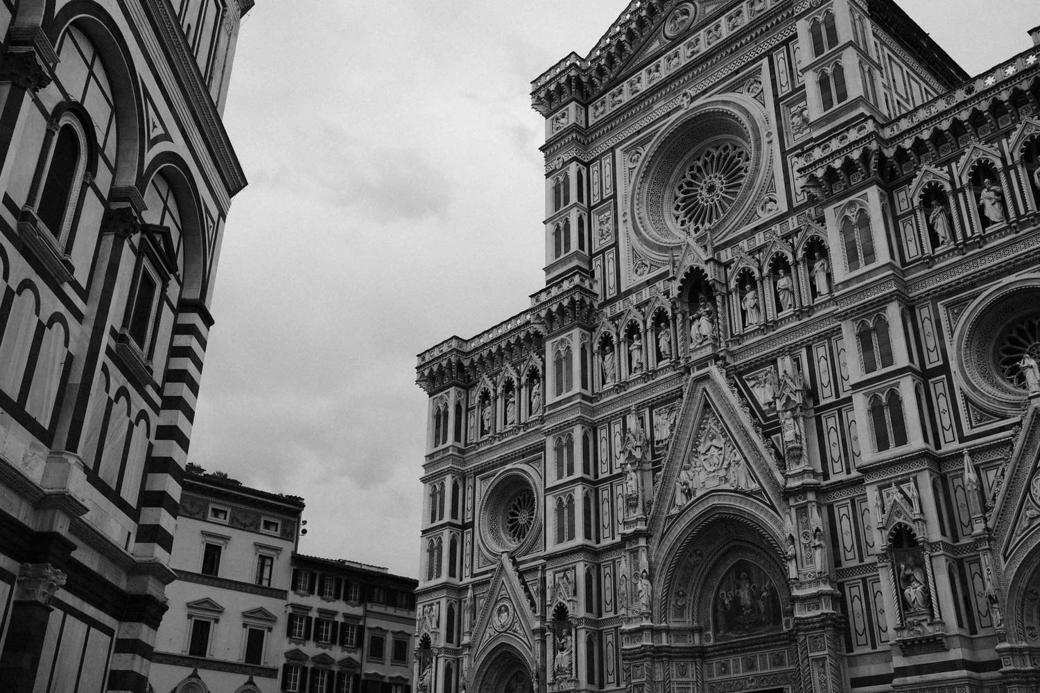 The ornate Gothic facade of Florence Cathedral rises dramatically against a cloudy sky in black and white.