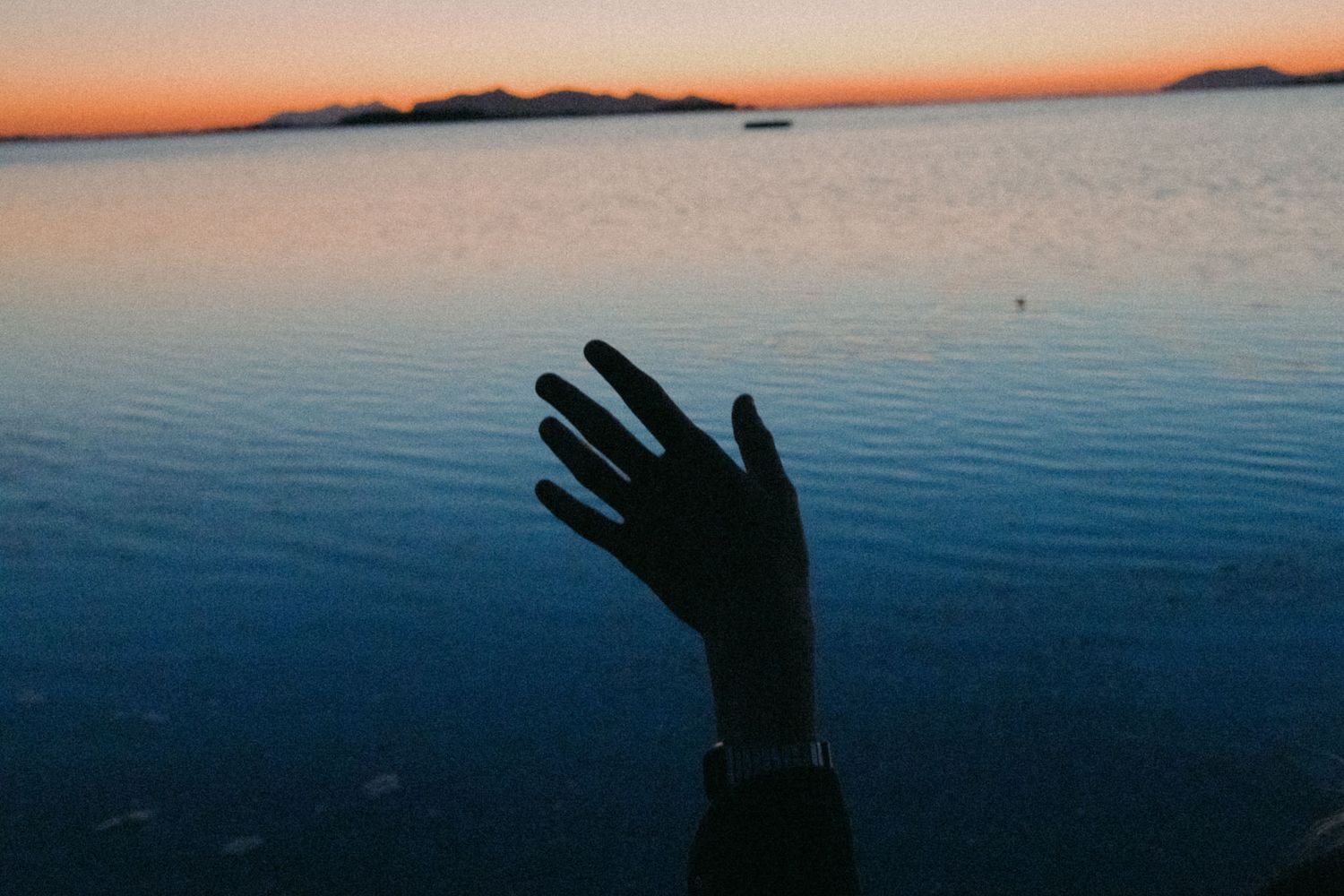A silhouetted hand reaches up against a serene sunset over calm water with mountains in the distance.