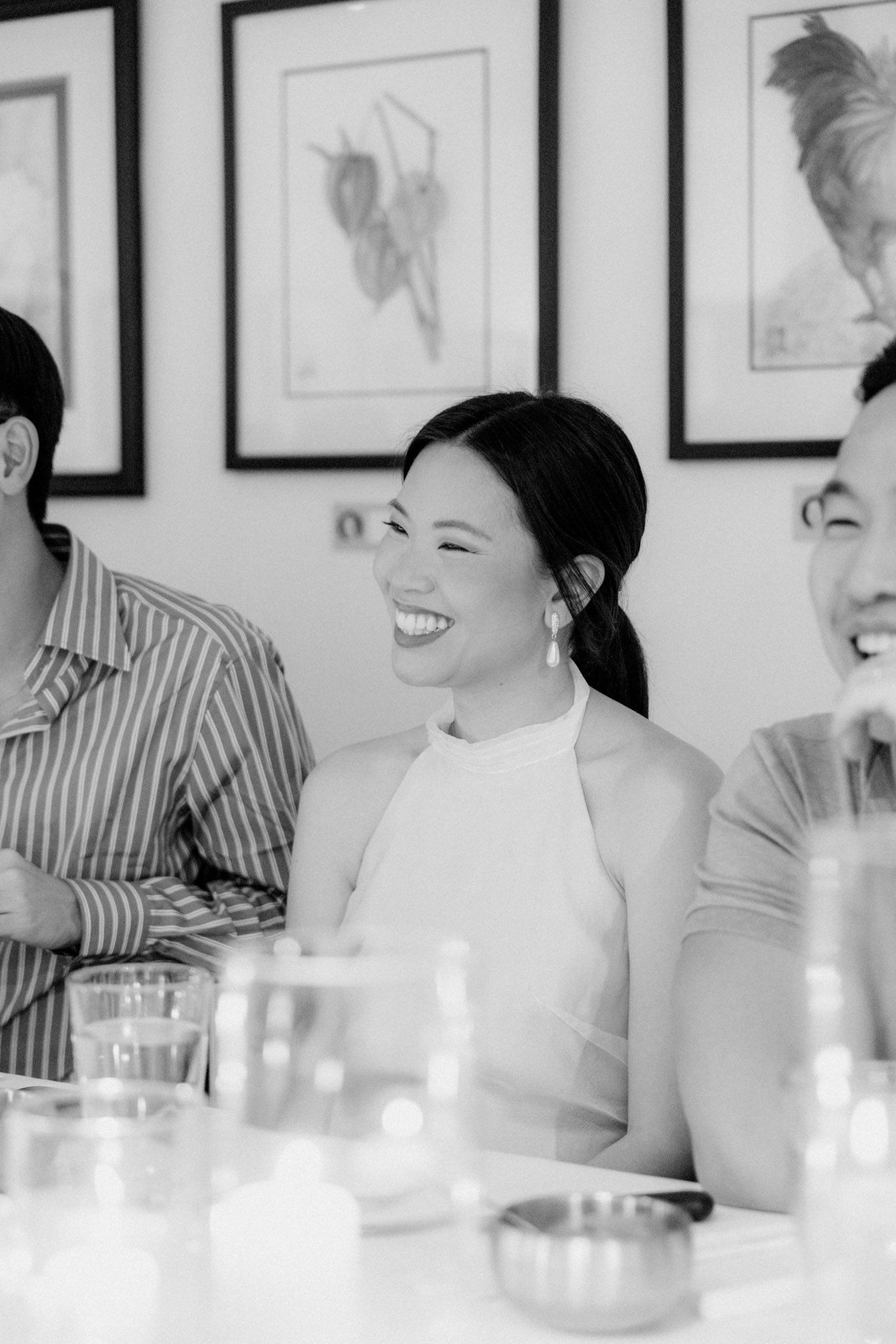 Black and white photo showing joyful laughter at a dinner table with framed artwork on the wall behind.