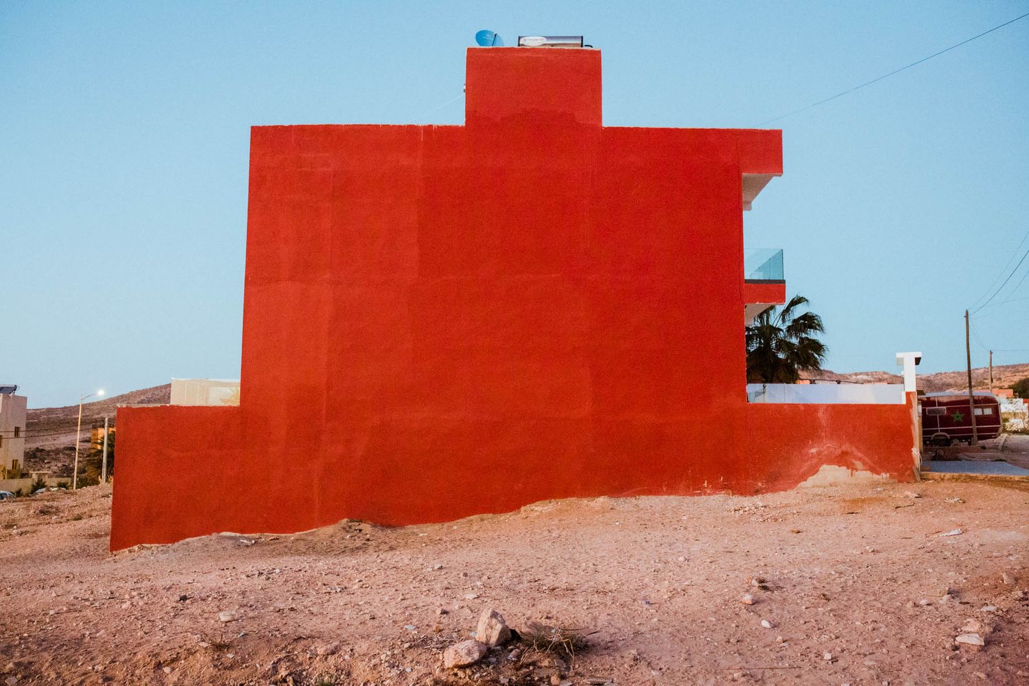Bright red painted building wall with a palm tree against clear blue sky in desert landscape.