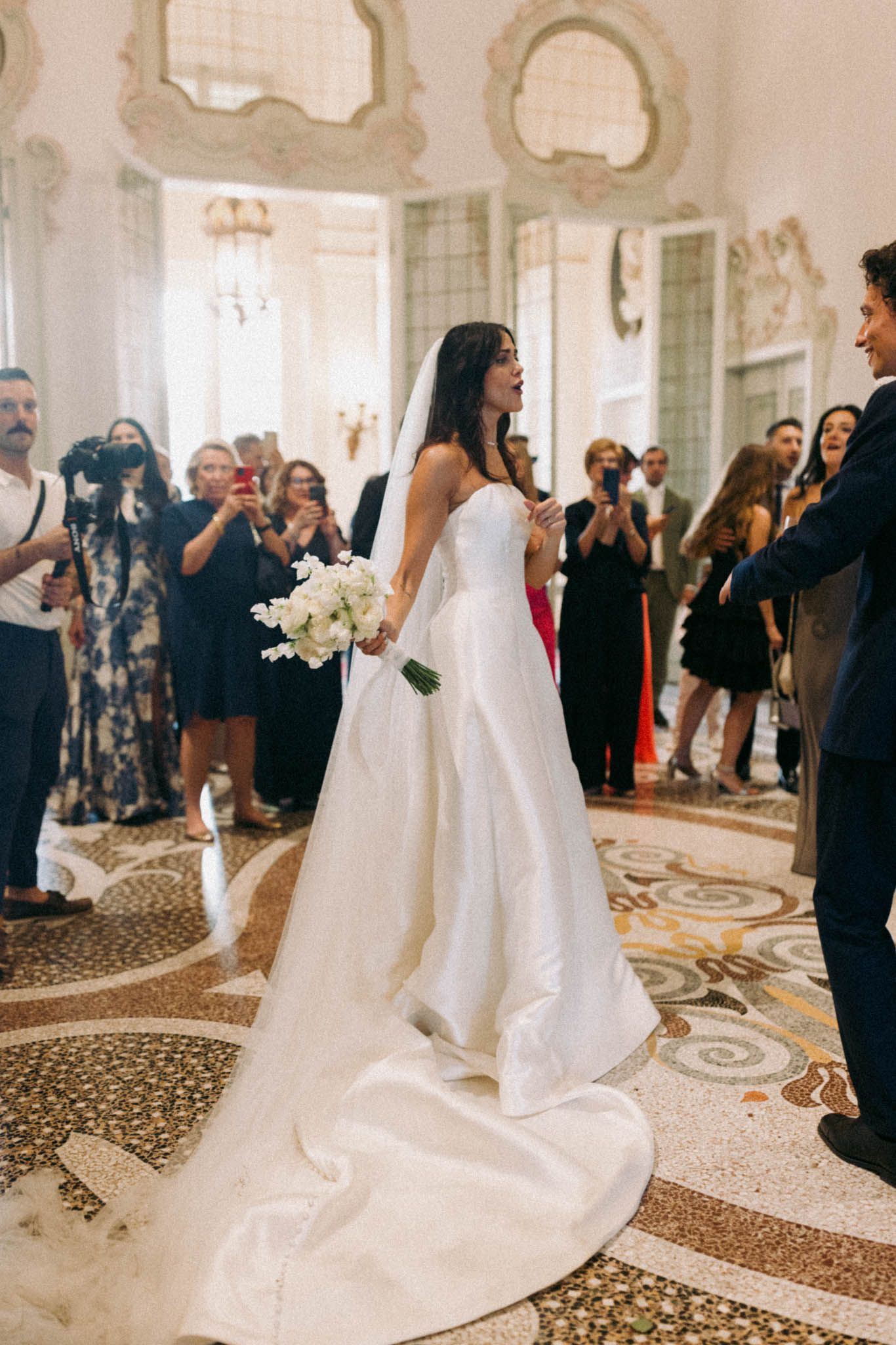 Wedding ceremony in an elegant ballroom with ornate carpeting and guests watching a bride in a flowing white gown.