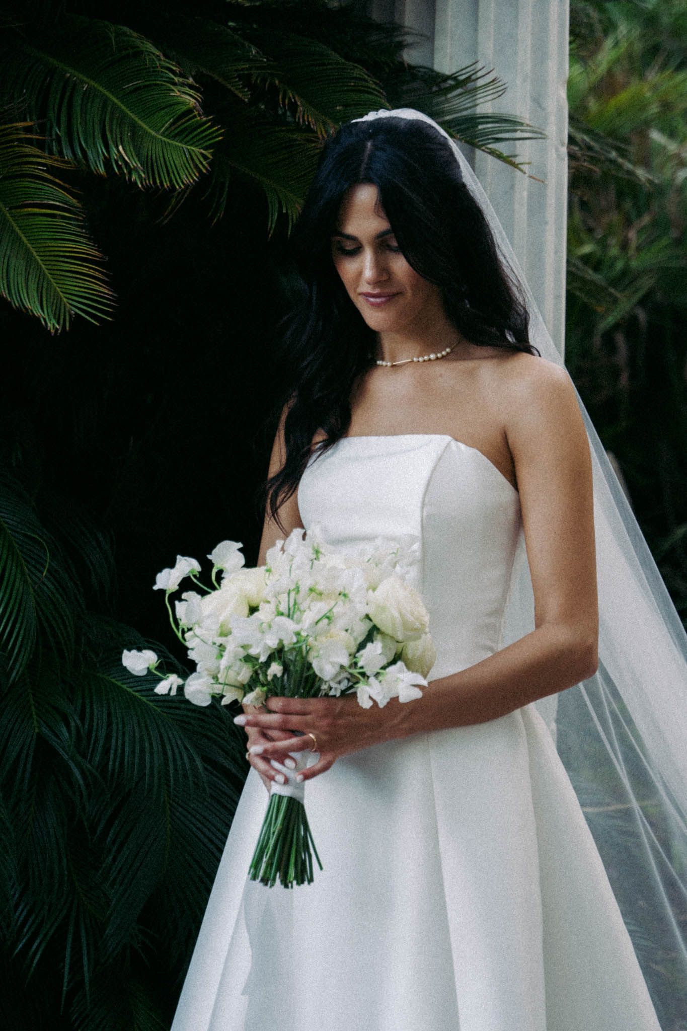 A bride in an elegant white strapless gown holds a beautiful bouquet of white roses and greenery.