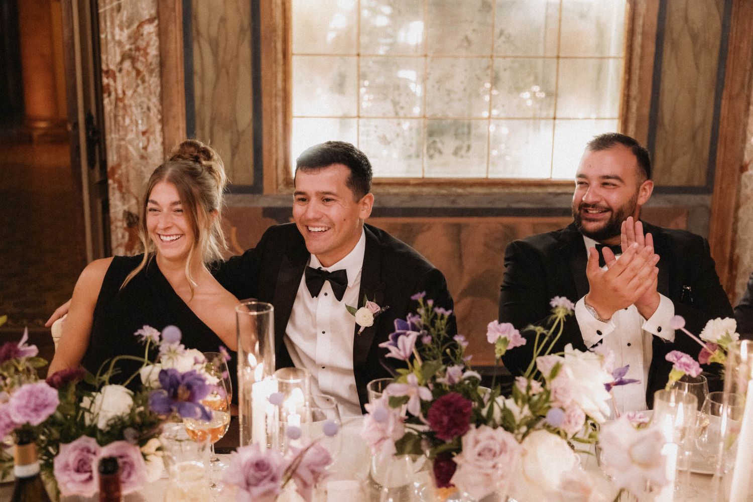 Wedding reception guests laugh and smile at a long candlelit table decorated with pink floral arrangements.