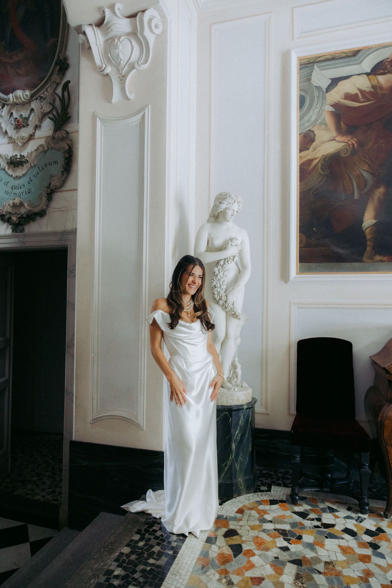 A person in a white dress poses against a white wall with ornate moldings and scattered autumn leaves on the floor.