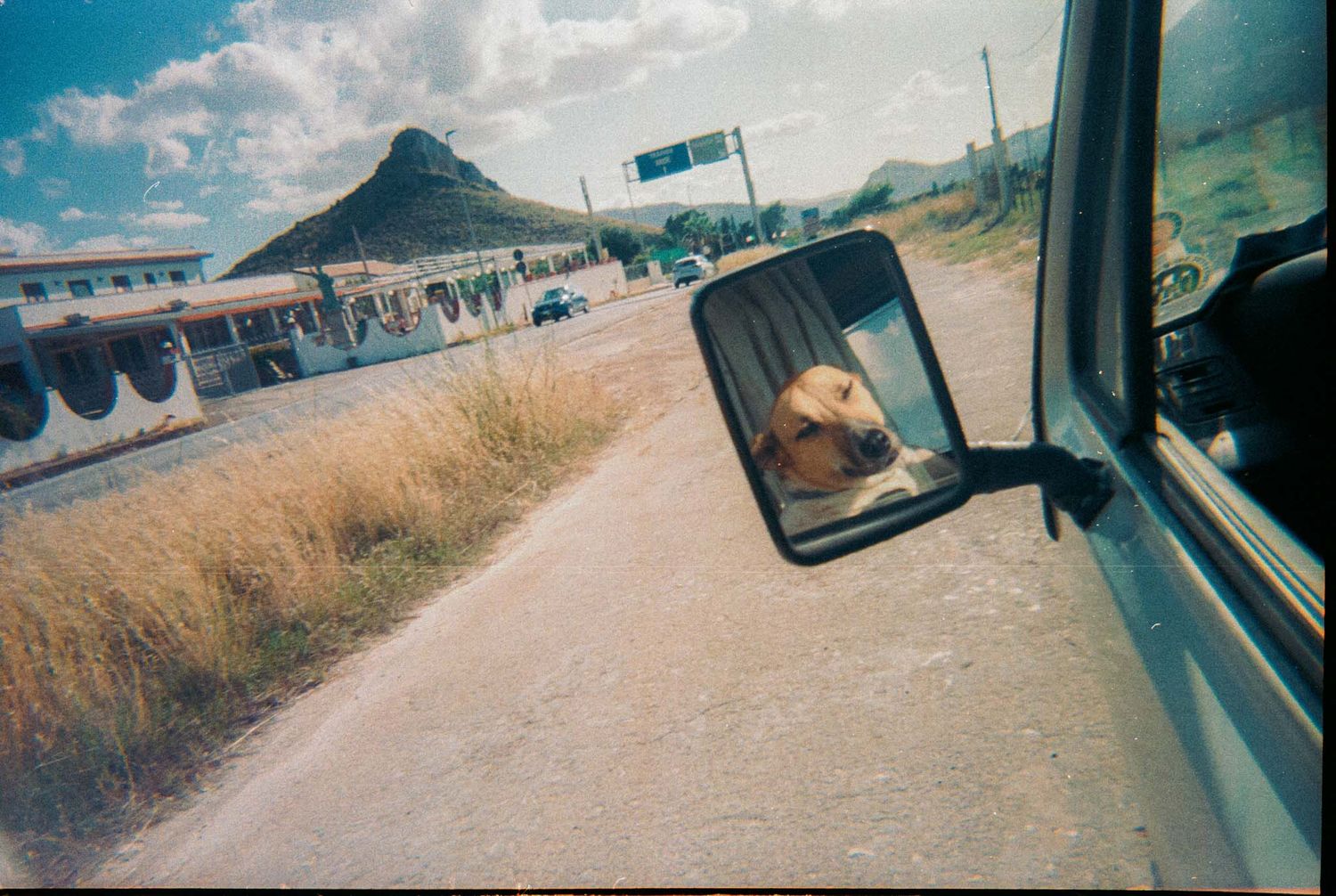 View from car side mirror showing desert road and mountain landscape with buildings in distance.