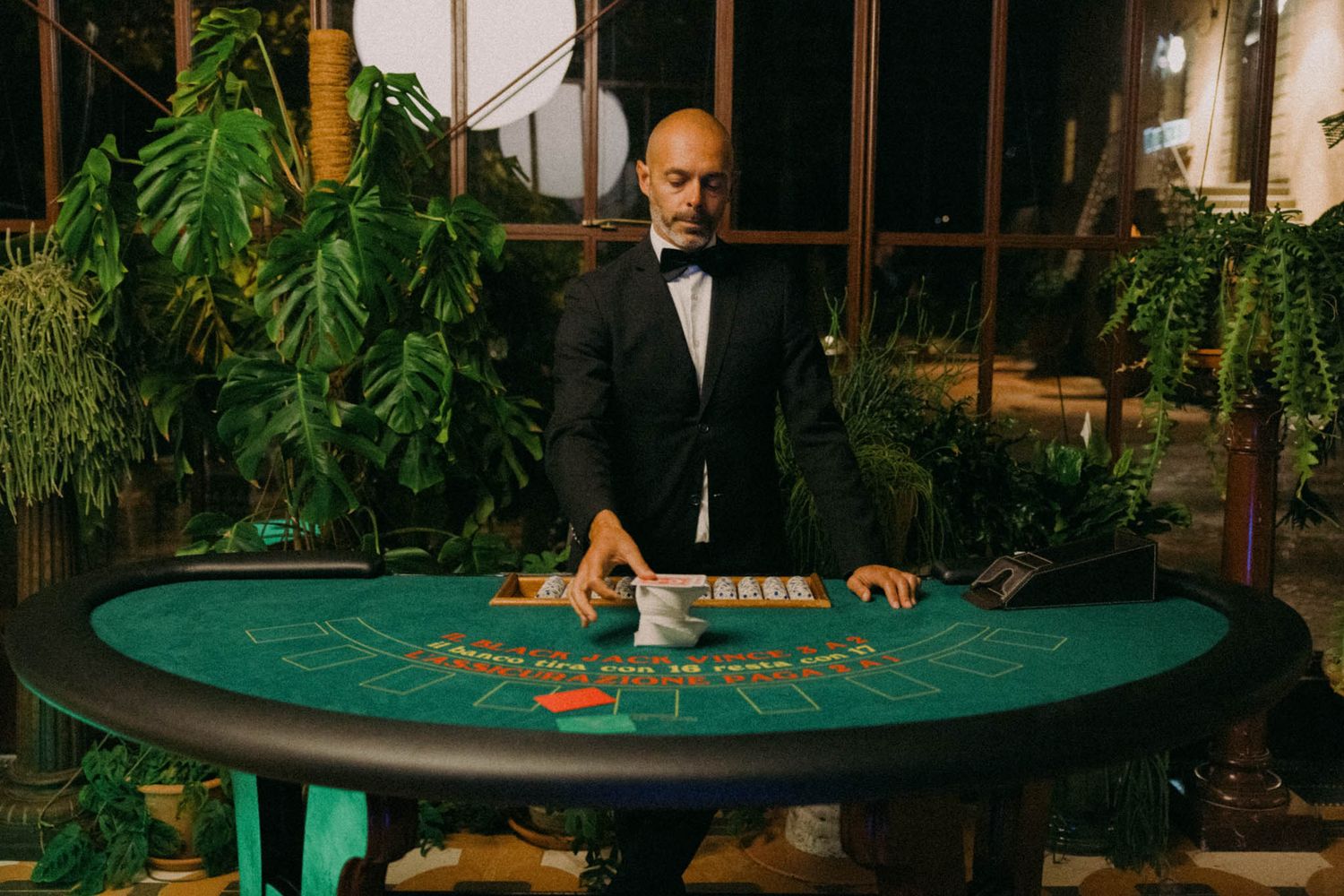 Casino dealer stands behind a green blackjack table surrounded by tropical plants in an elegant setting.