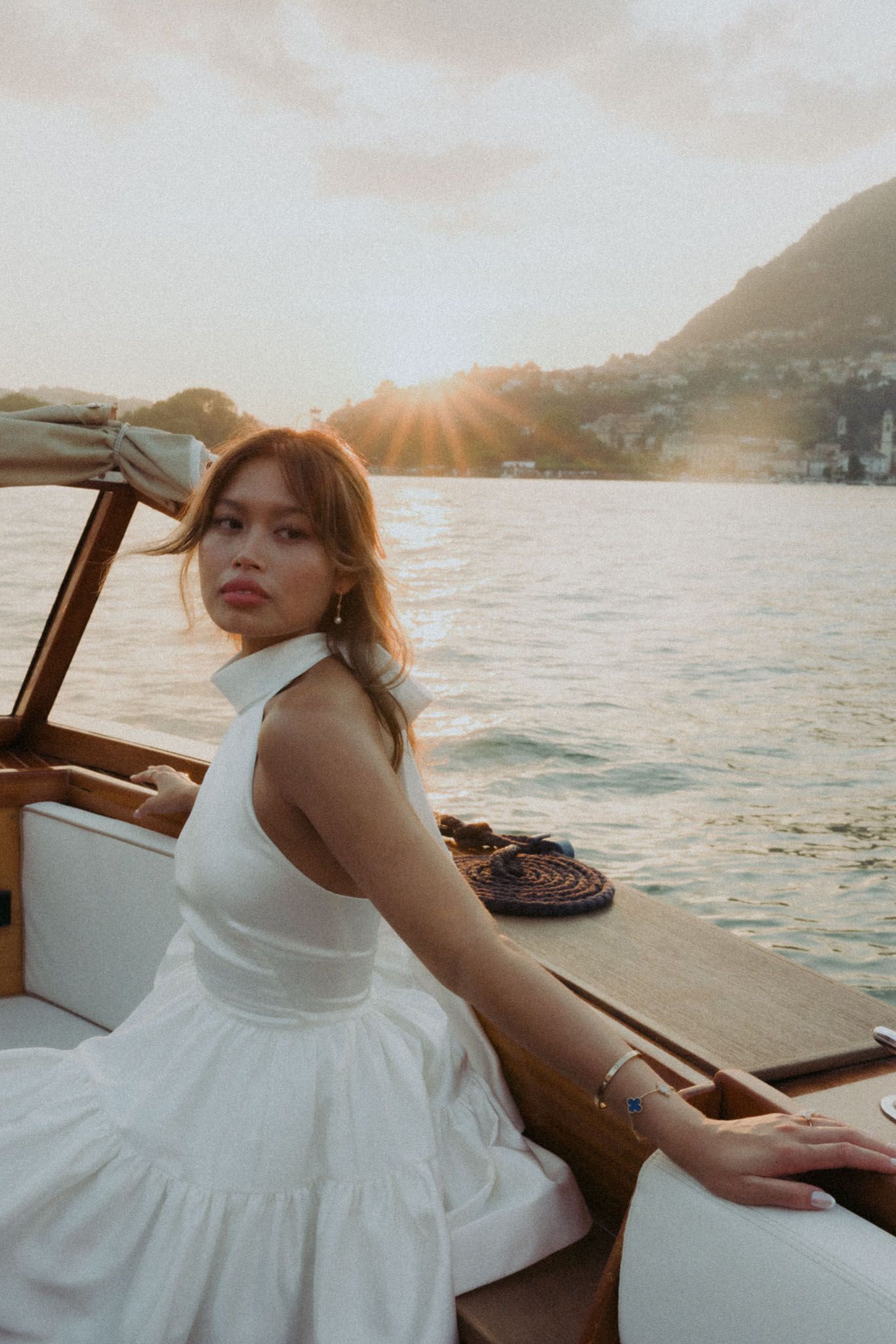 A person in a white dress sits on a boat at sunset with mountains and water in the background.