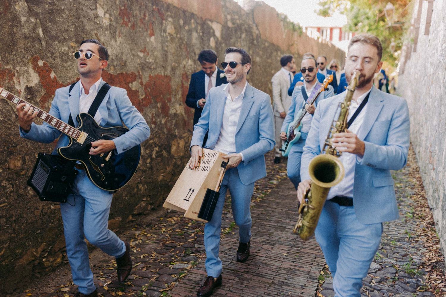 Musicians in matching light blue suits perform with guitar, percussion and saxophone while walking down a stone path.