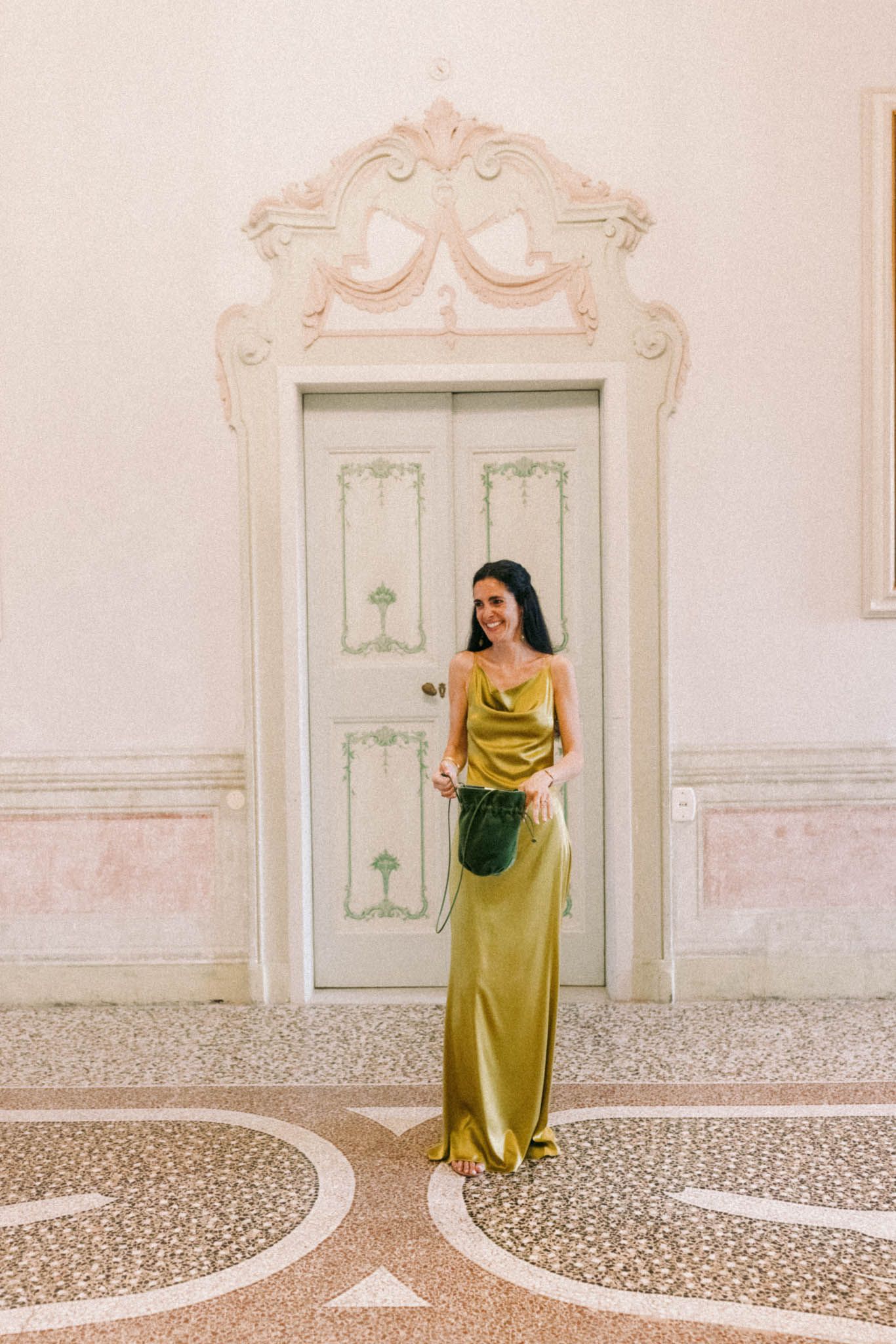 A figure in a chartreuse gown stands against ornate white walls and patterned tile floors in an elegant interior space.