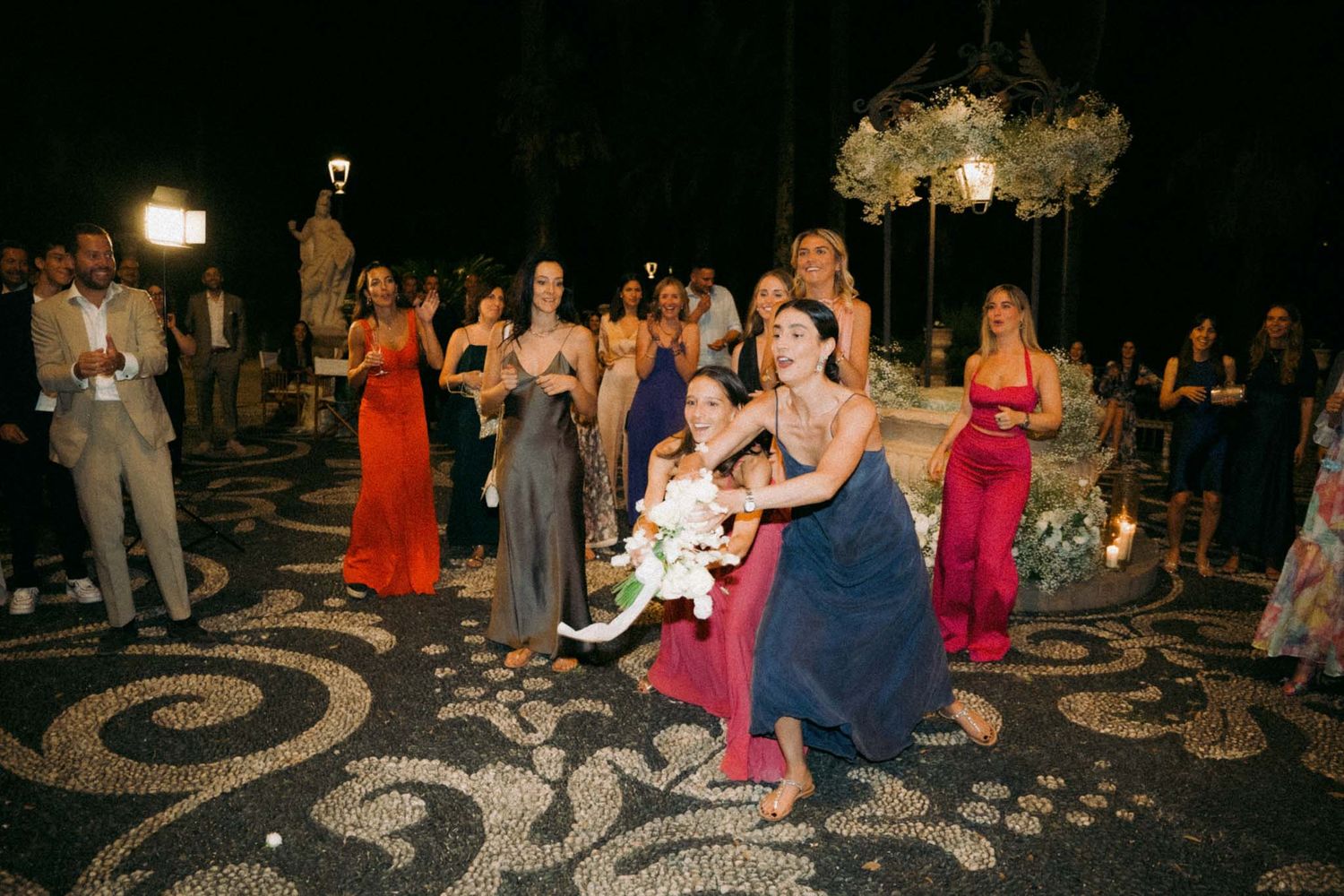 Wedding guests gather on an ornate mosaic patio at night during the bouquet toss tradition.