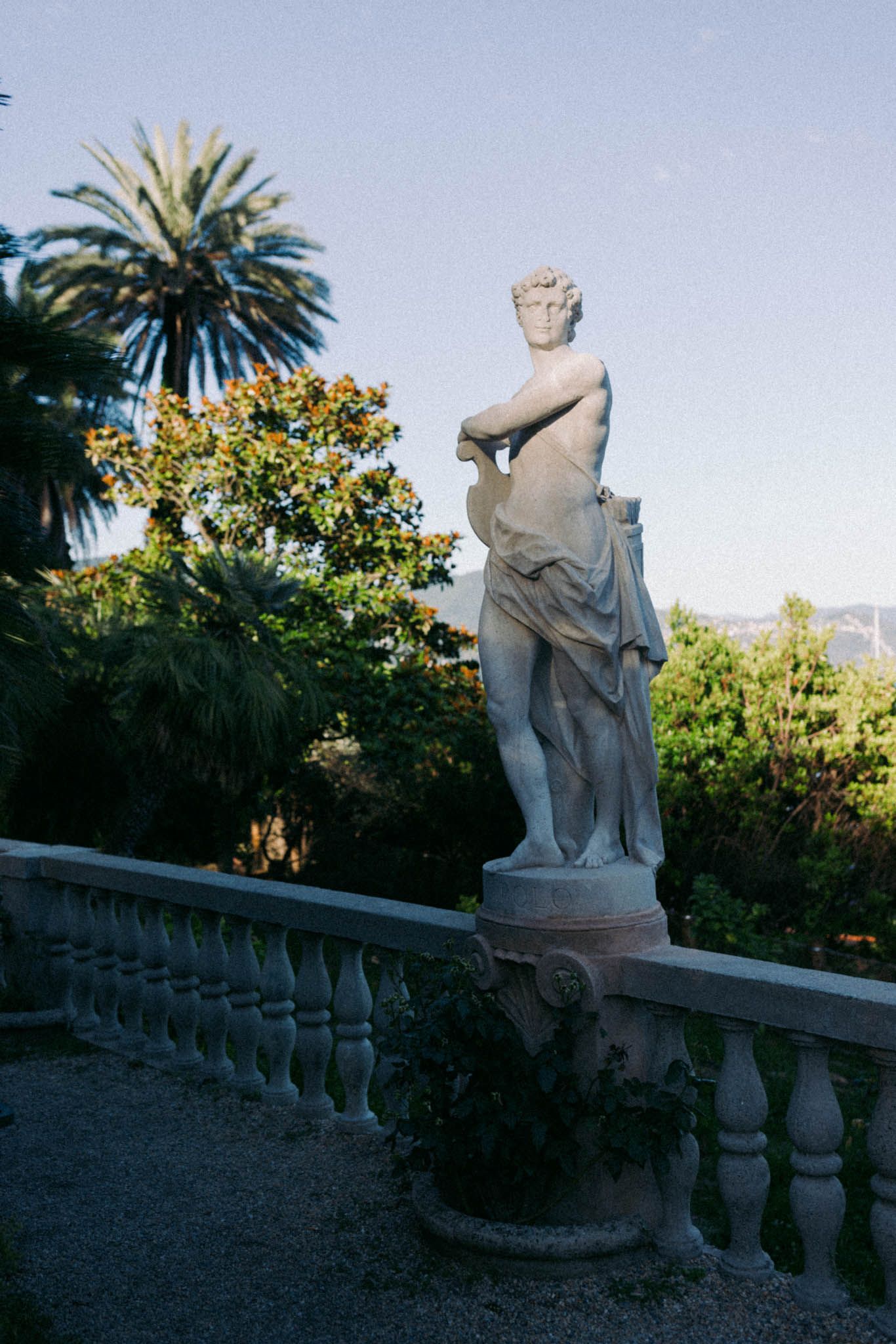 Classical marble statues line a garden terrace with palm trees and lush greenery in the background.