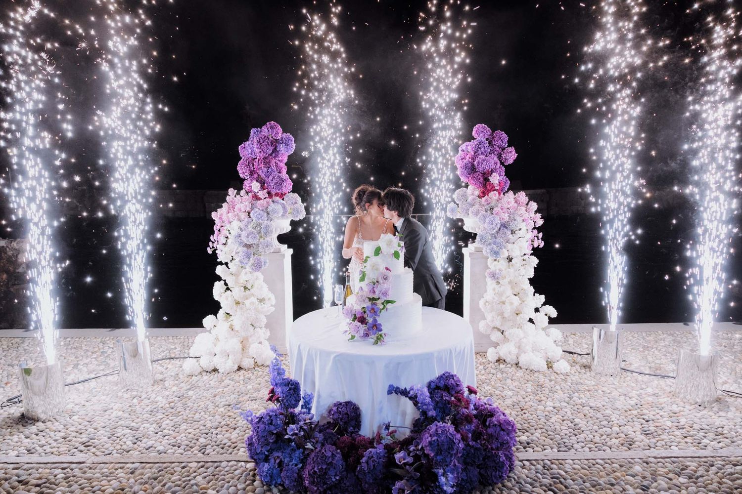 White tiered cake adorned with purple flowers surrounded by sparkling fountains at an evening reception.
