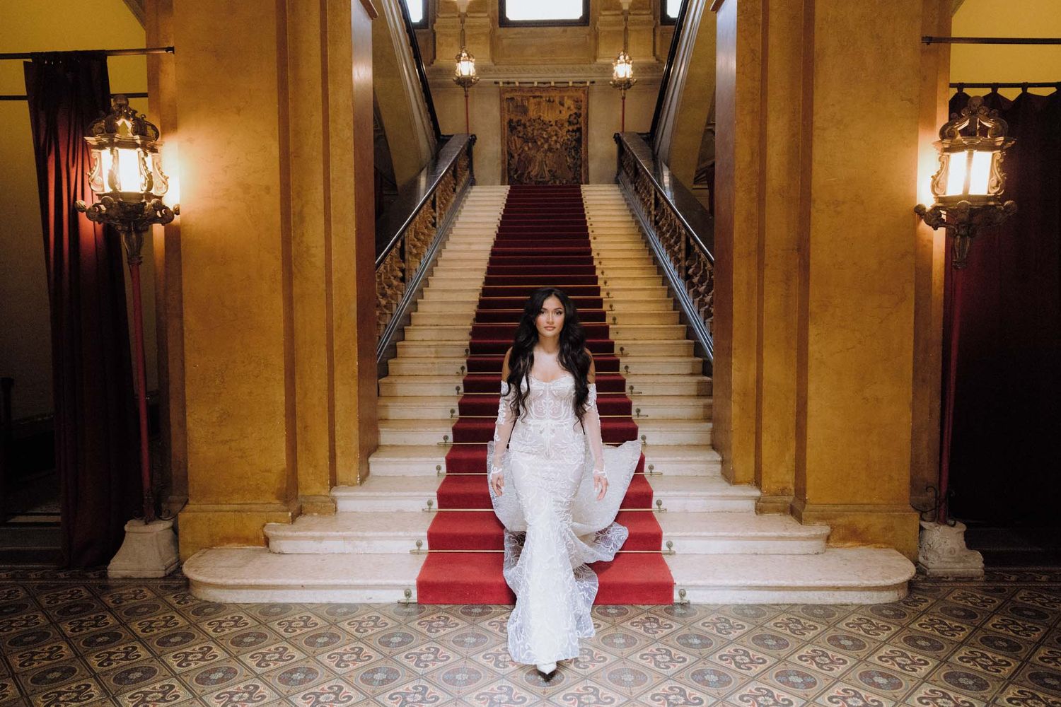 A person in a white dress walks up an elegant staircase with red carpet and ornate golden walls.