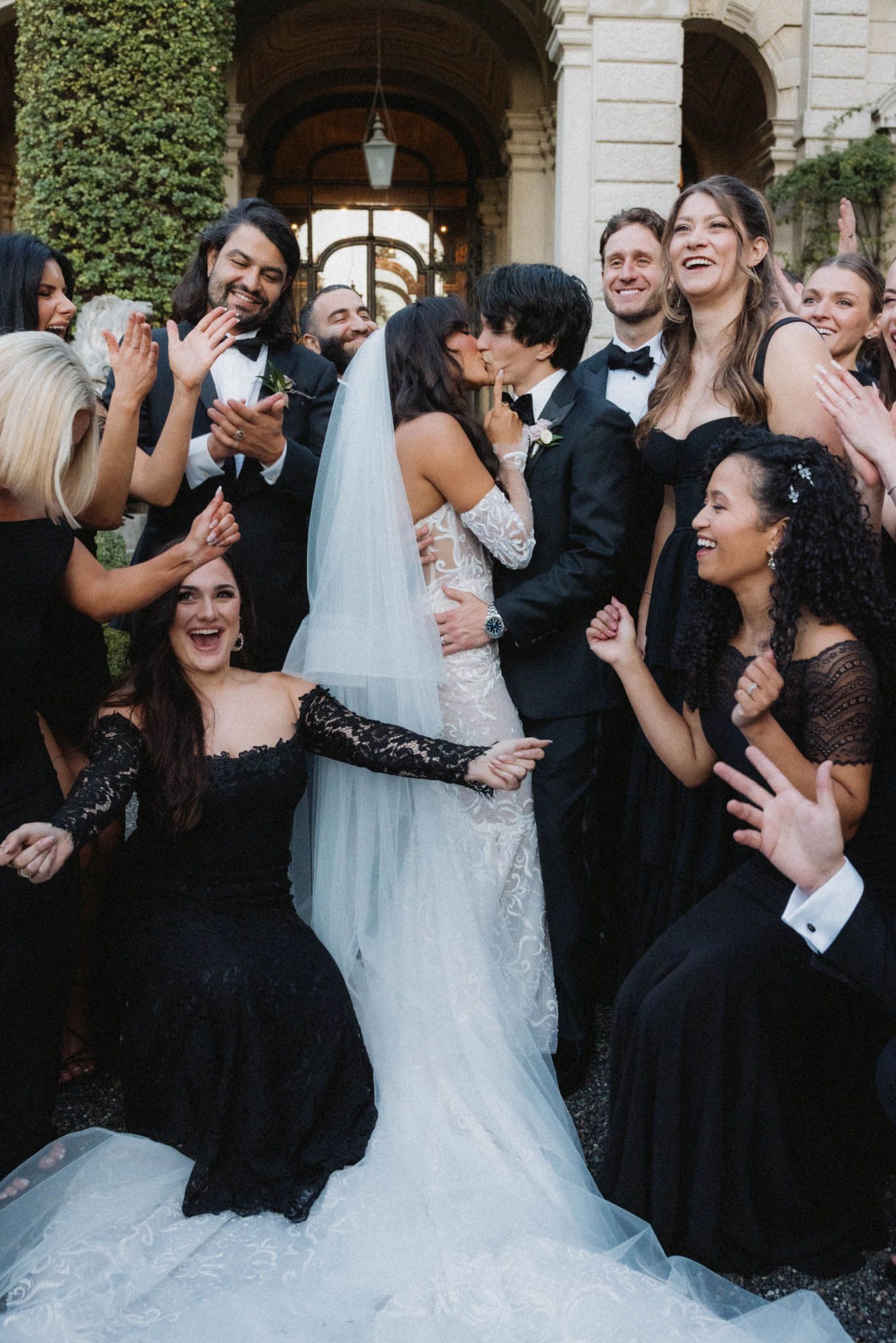 Wedding party guests celebrate and laugh together in formal attire at an outdoor venue with ivy-covered walls.