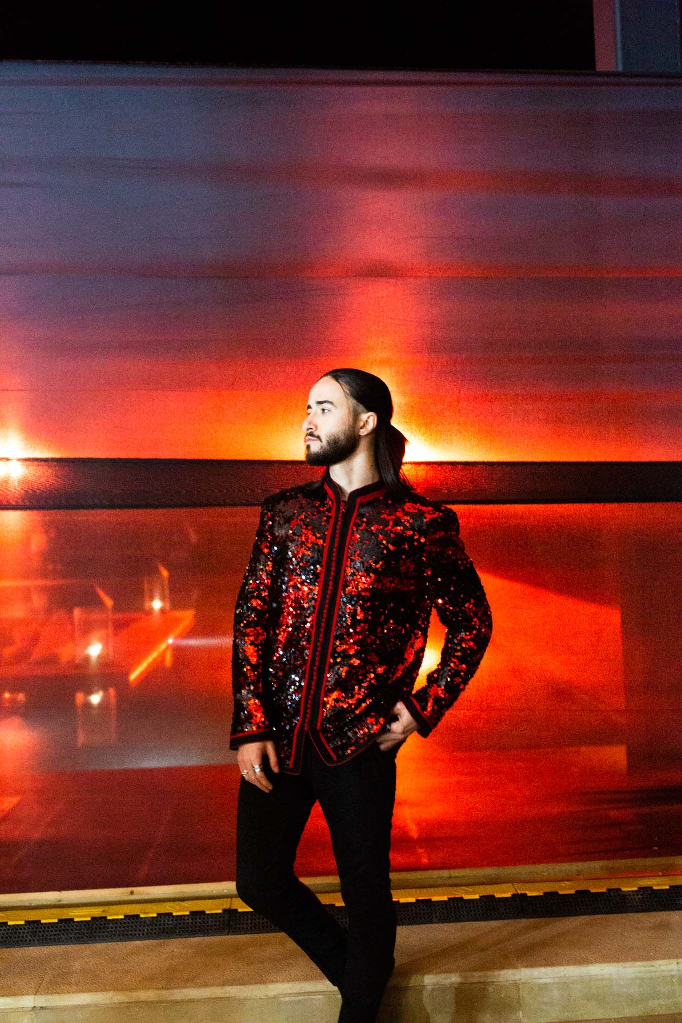 A person in a sequined jacket poses against a dramatic red sunset backdrop creating a striking silhouette effect.